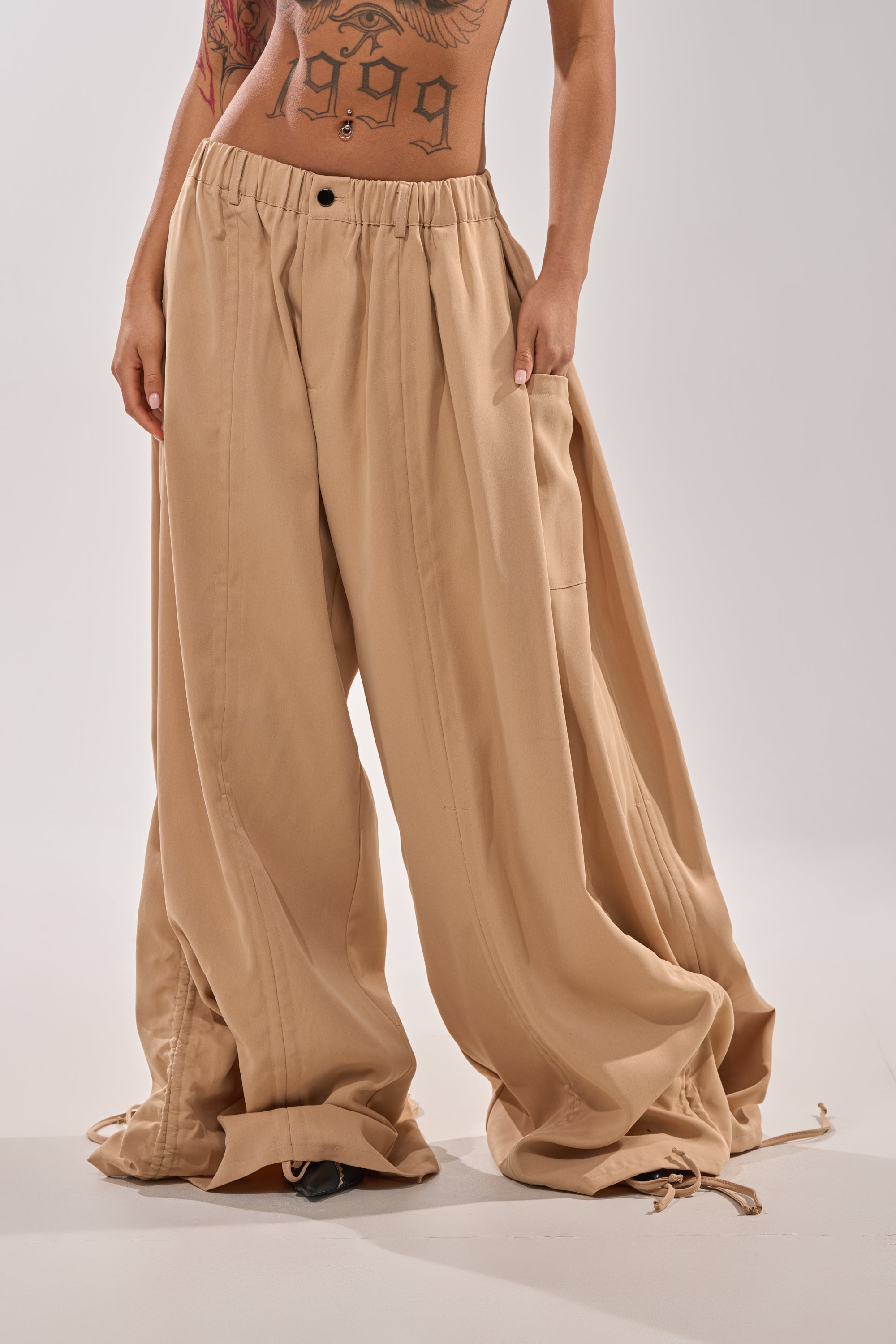 A person models the DRAMA RUCHED JOGGER IN KHAKI, featuring a high-rise elastic waistband, against a plain light background. Their face is hidden, spotlighting the street style look and ruched details of these khaki joggers. Tattoos are visible on their torso and arms. 4
