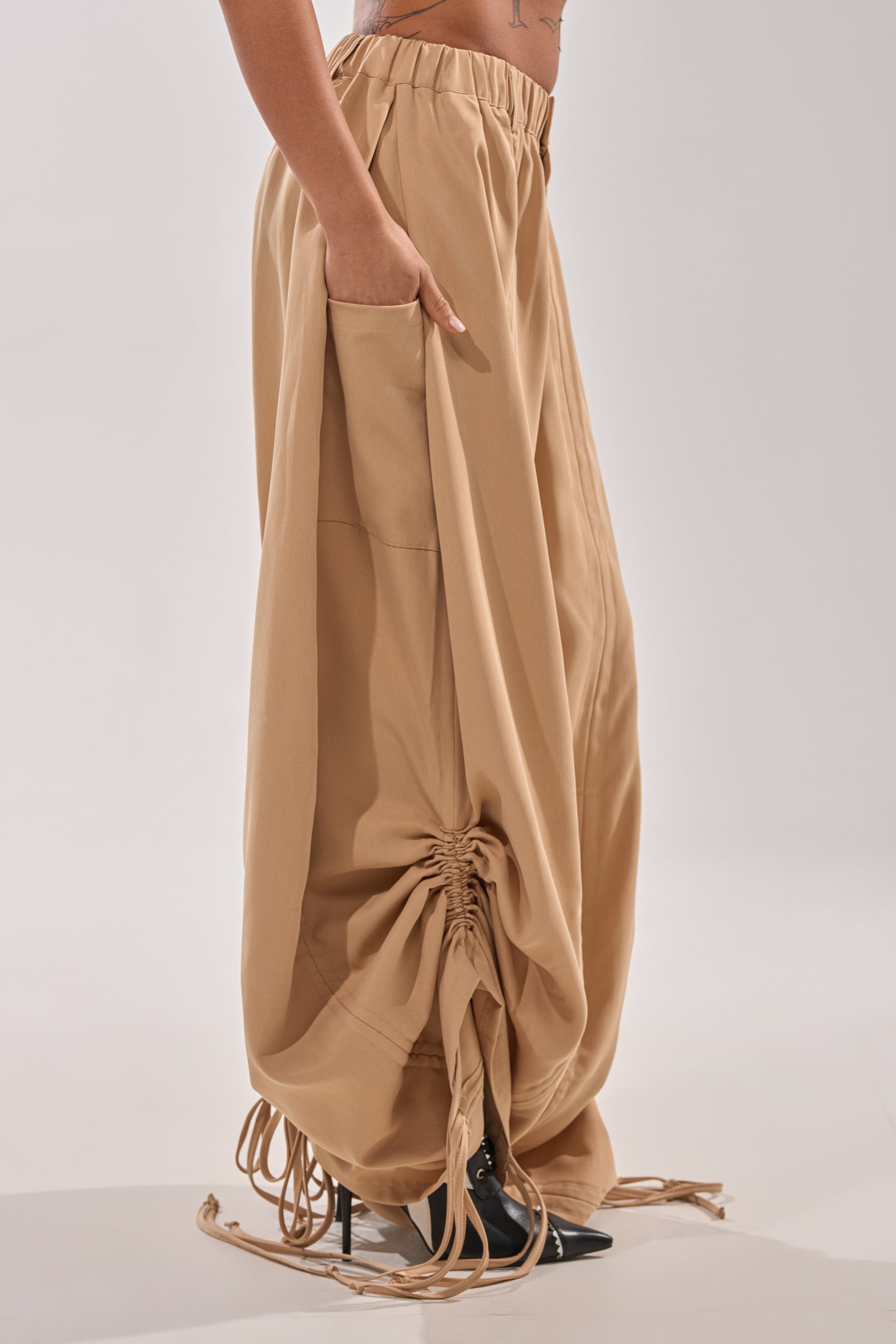 A person is shown from the side against a plain background wearing the DRAMA RUCHED JOGGER IN KHAKI, featuring a high-rise elastic waistband, ruched detailing, and side pockets, styled with high heels. 5