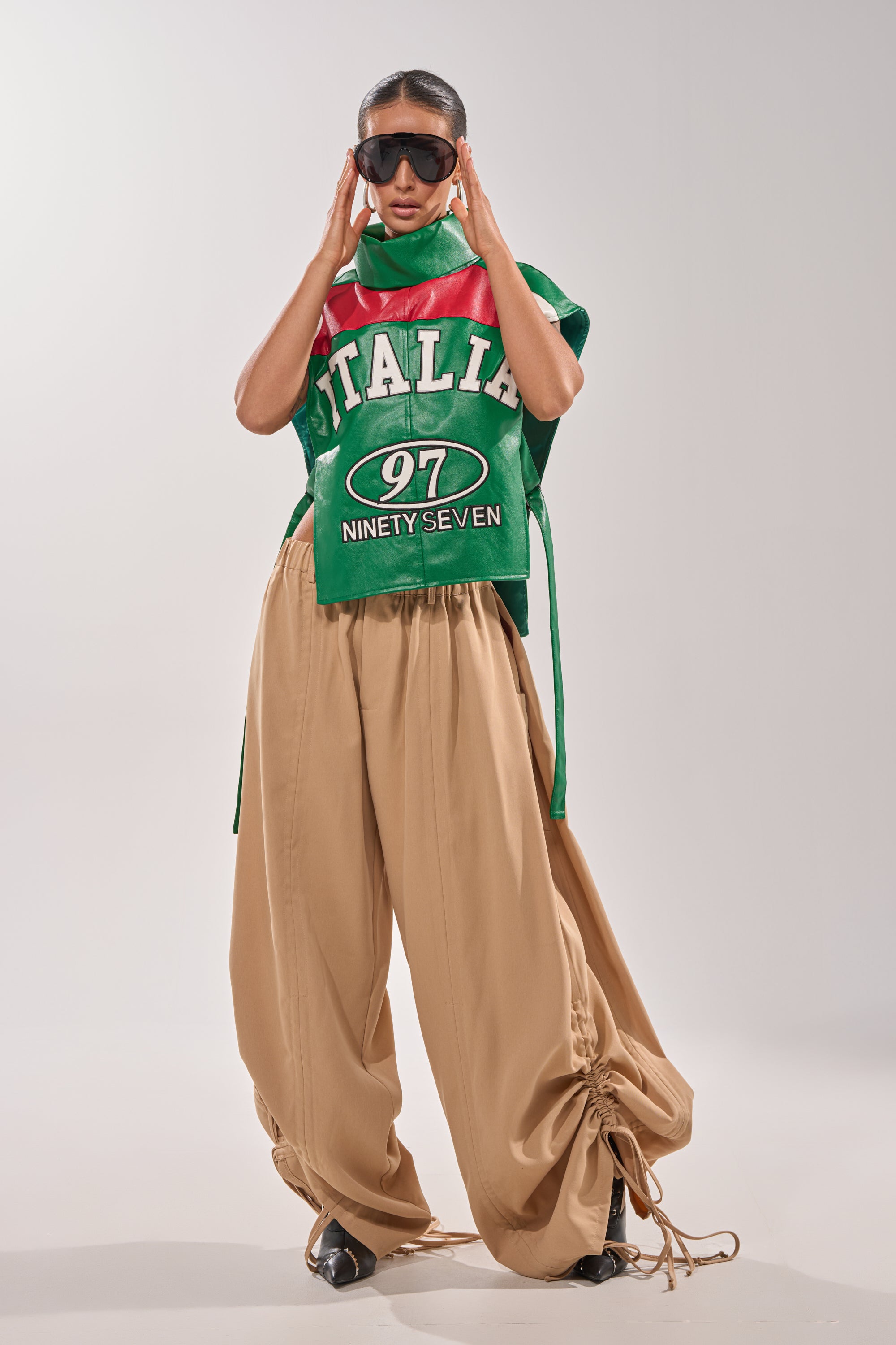 A person poses confidently against a plain light background wearing DRAMA RUCHED JOGGER IN KHAKI, a green "ITALIA 97 NINETY SEVEN" sleeveless top, black shoes, and oversized black sunglasses. 7