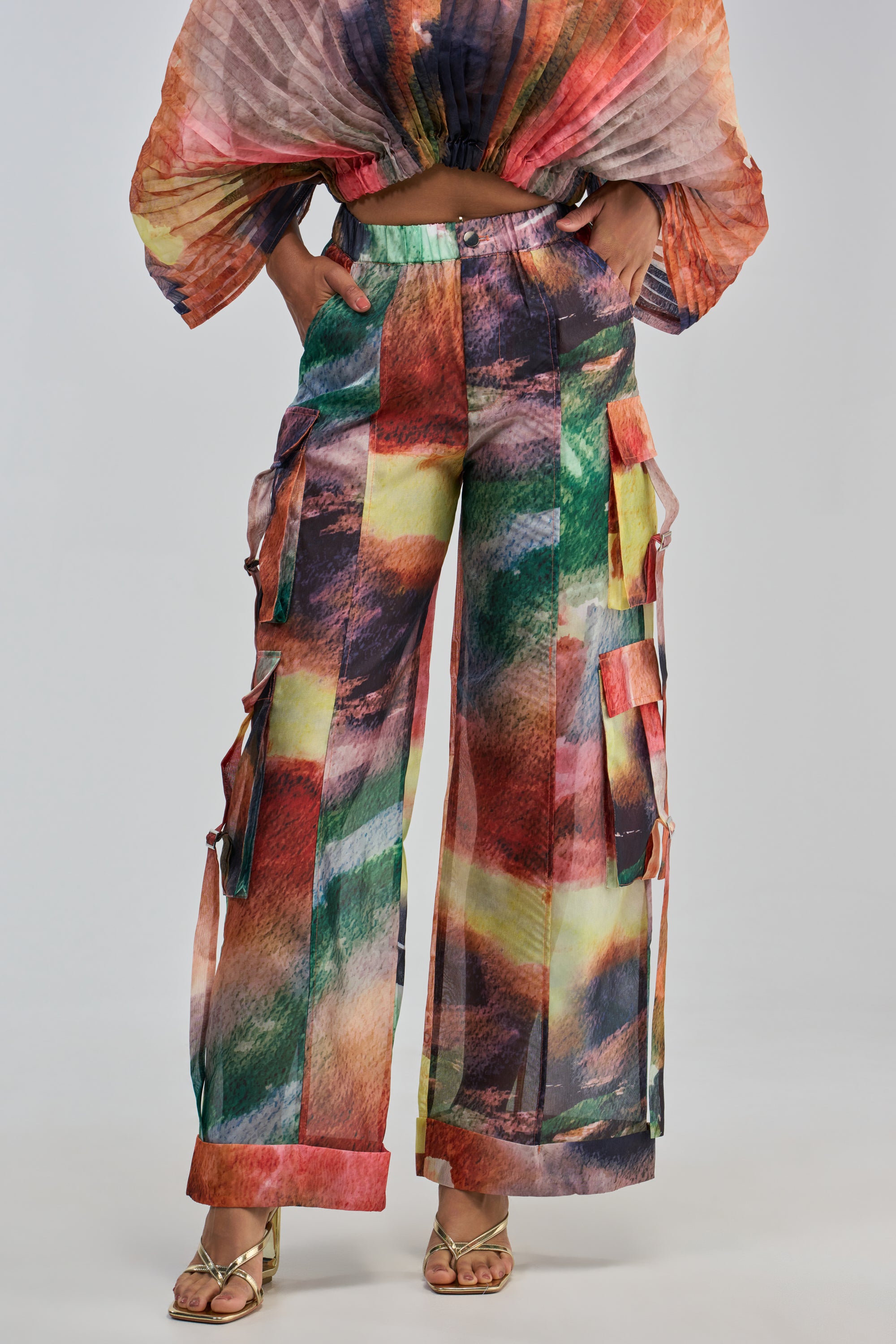 A woman stands against a plain gray background with her hands in the pockets of the Watercolors Chiffon Cargo Pant, a loose, high-waisted pant featuring a watercolor print and side pockets. She pairs it with a matching long-sleeve top and gold heels.