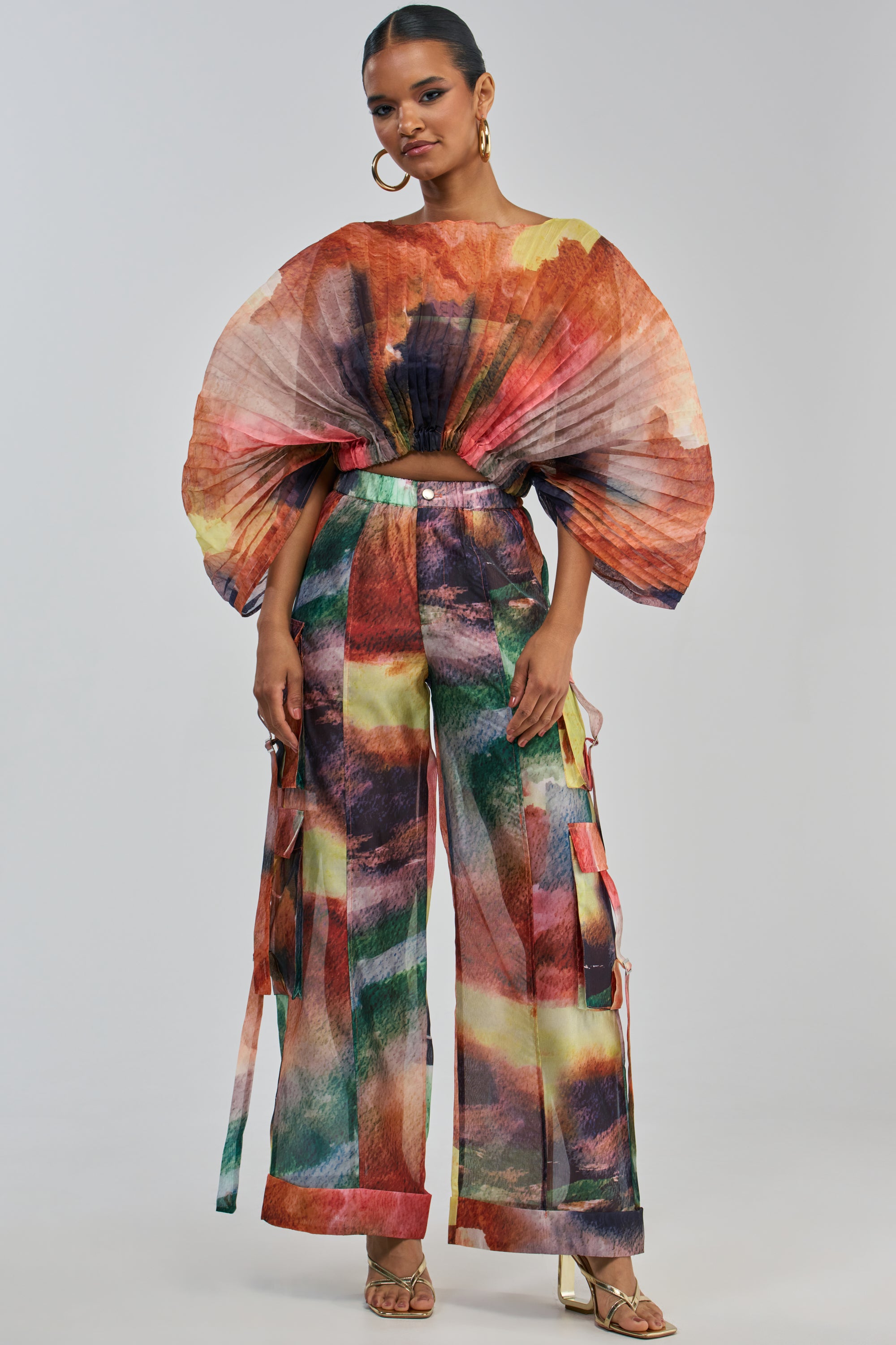 A woman wears the WATERCOLORS CHIFFON CARGO PANT, a vibrant piece featuring a bold abstract watercolor print. Paired with oversized pleated sleeves, heeled sandals, and large hoop earrings, she stands confidently against a plain background, embodying modern wide-leg streetwear style.