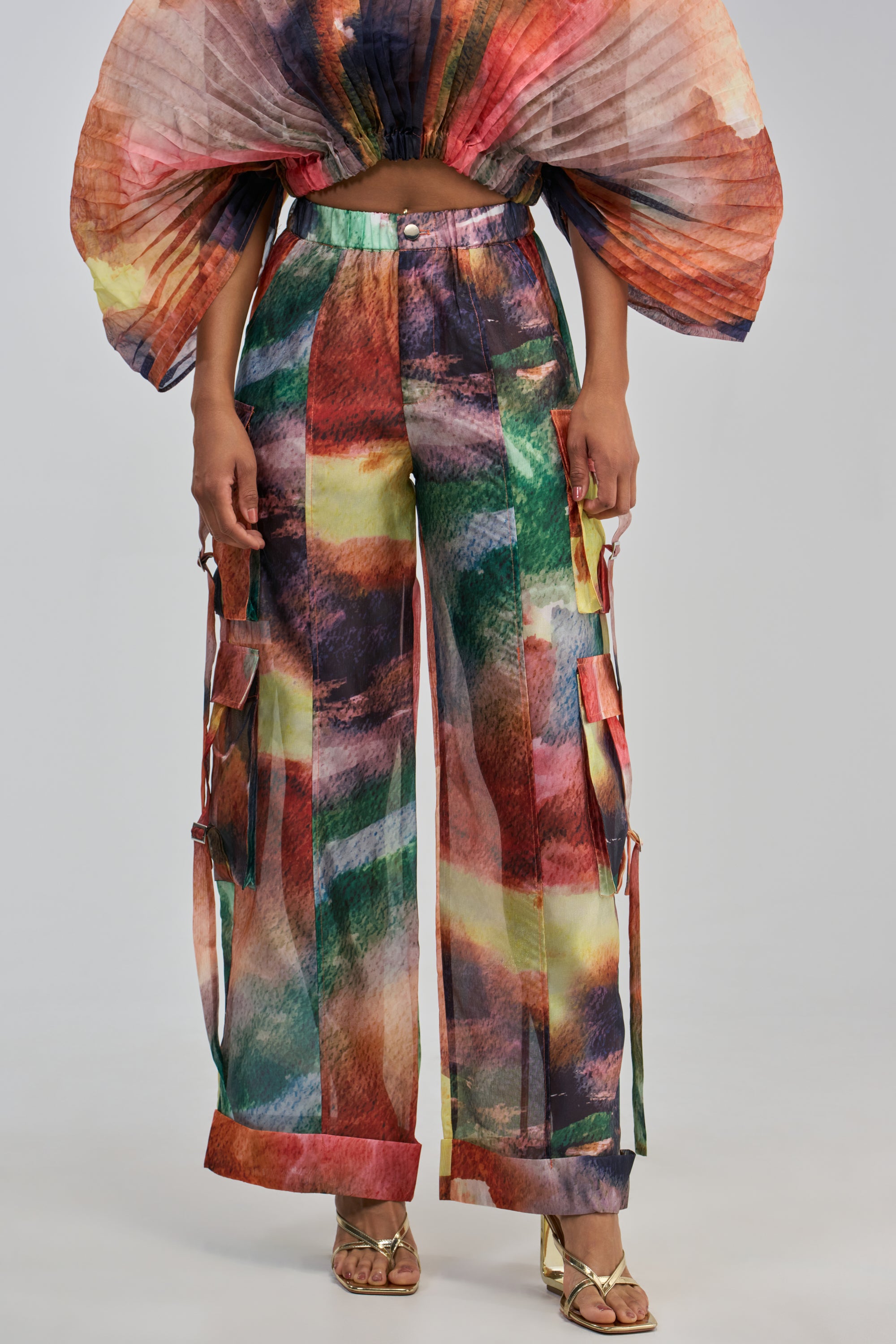 A model wears the WATERCOLORS CHIFFON CARGO PANT, featuring vibrant abstract patterns in red, green, yellow, and purple. The pants are styled with a matching pleated, wing-like top and gold sandals for a colorful streetwear look.
