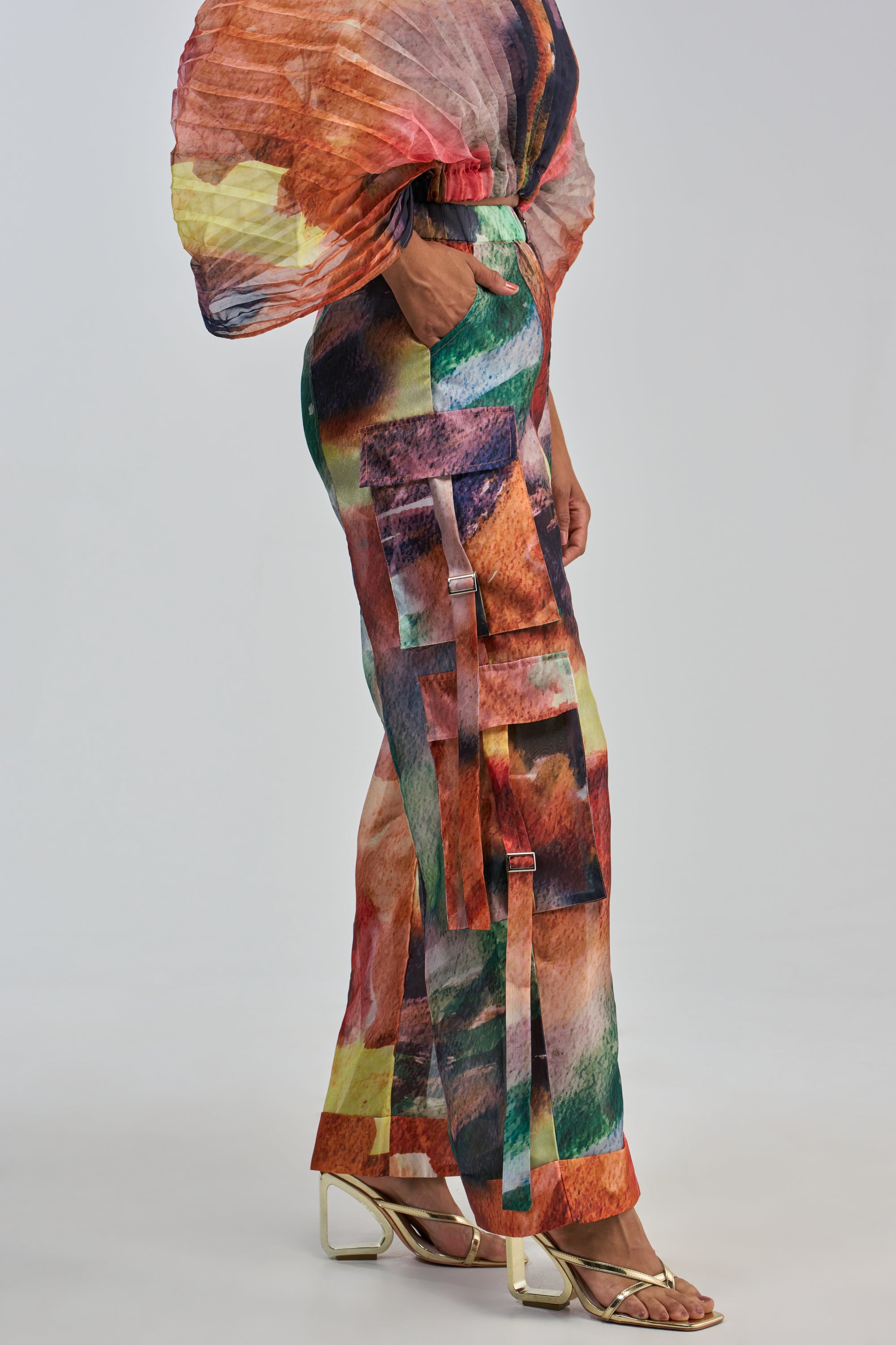 Dressed in the WATERCOLORS CHIFFON CARGO PANT and a coordinating top with puffy sleeves, a person stands against a plain background, completing the look with gold open-toe heels.