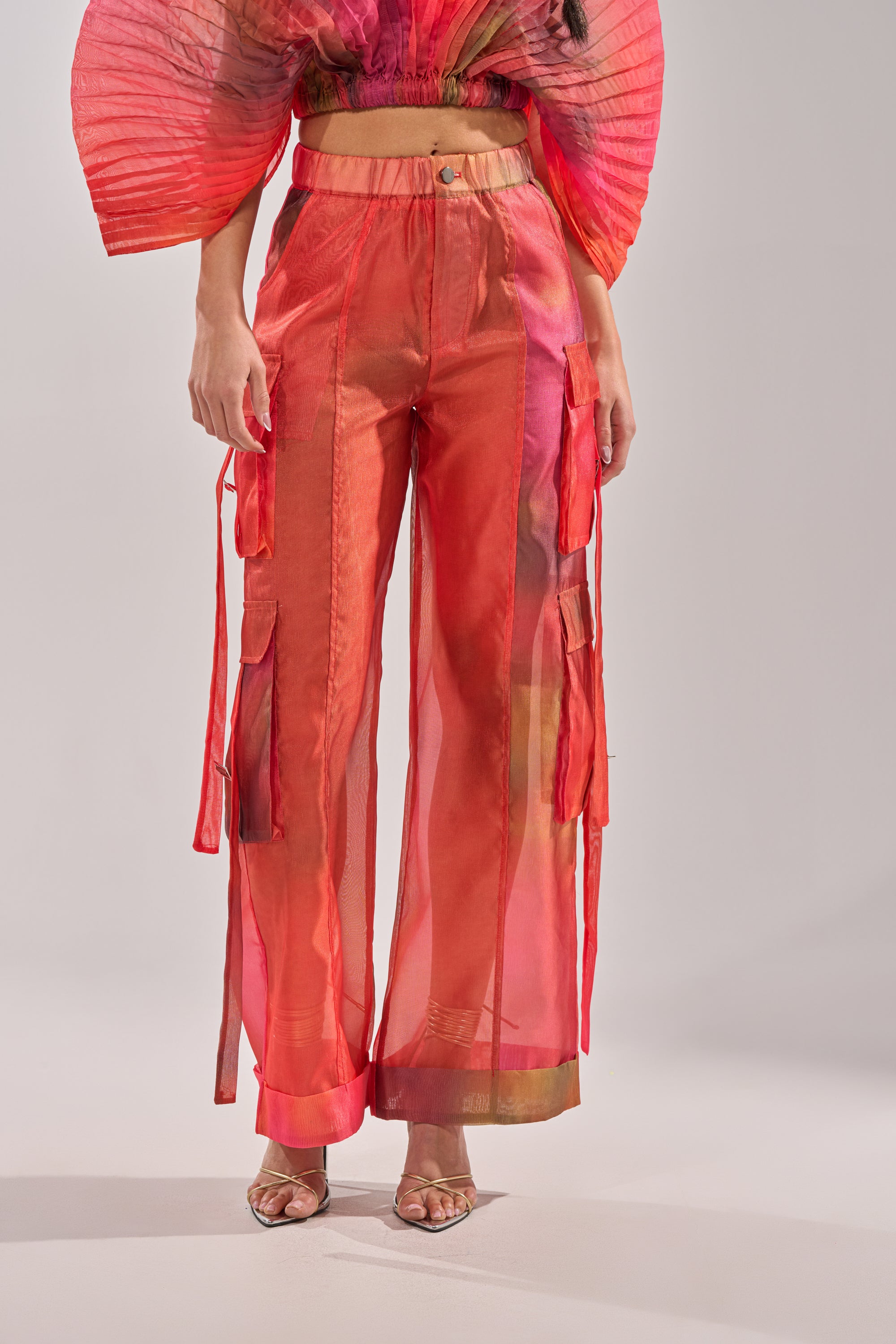 A person wears the STAINED RED LIGHTWEIGHT CARGO PANT, featuring sheer, wide-leg chiffon and cargo pockets with ribbon details, paired with a matching crop top with dramatic pleated sleeves and open-toe heels. 1