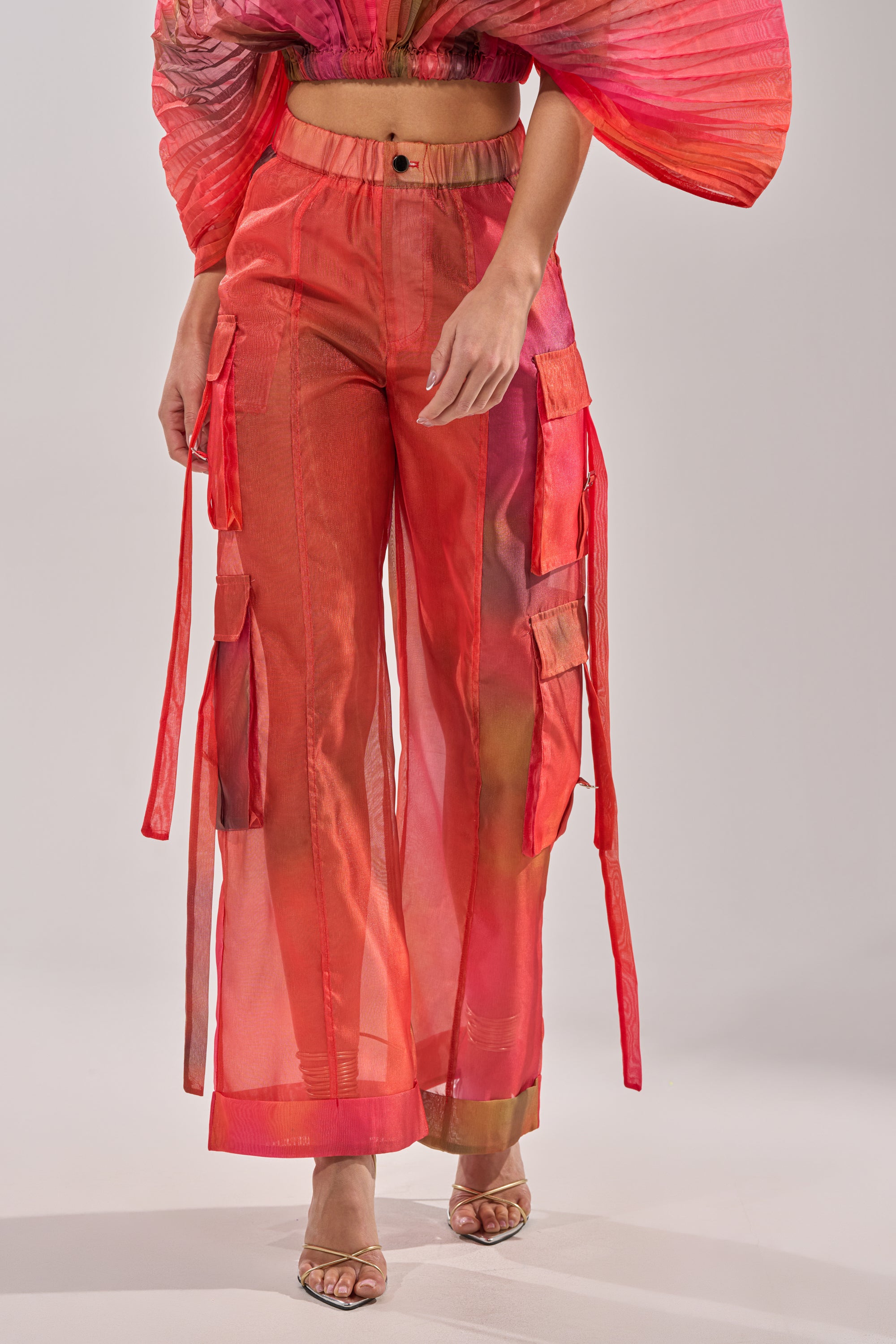 A person models the STAINED RED LIGHTWEIGHT CARGO PANT in a sheer, bright red wide-leg silhouette with pockets, paired with a matching cropped top with pleated sleeves and open-toed heels against a plain, light background. 2