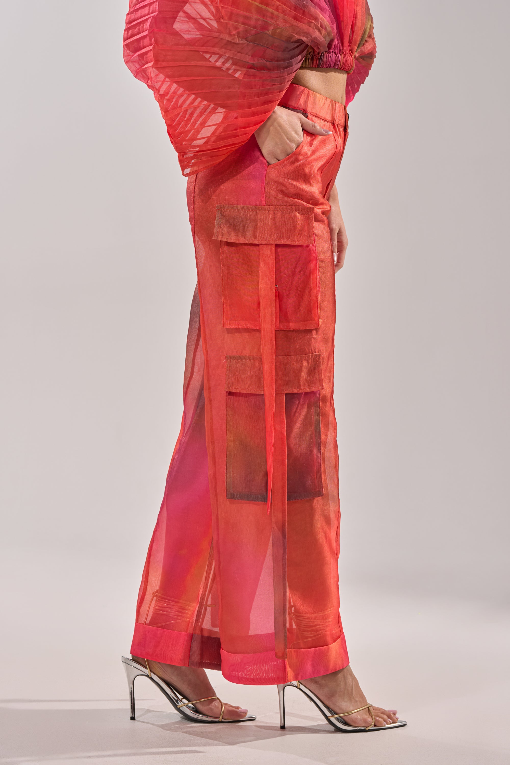 Wearing the STAINED RED LIGHTWEIGHT CARGO PANT, featuring a sheer fabric, large pockets, and a matching top, someone poses against a plain light background with silver high-heeled sandals. 3