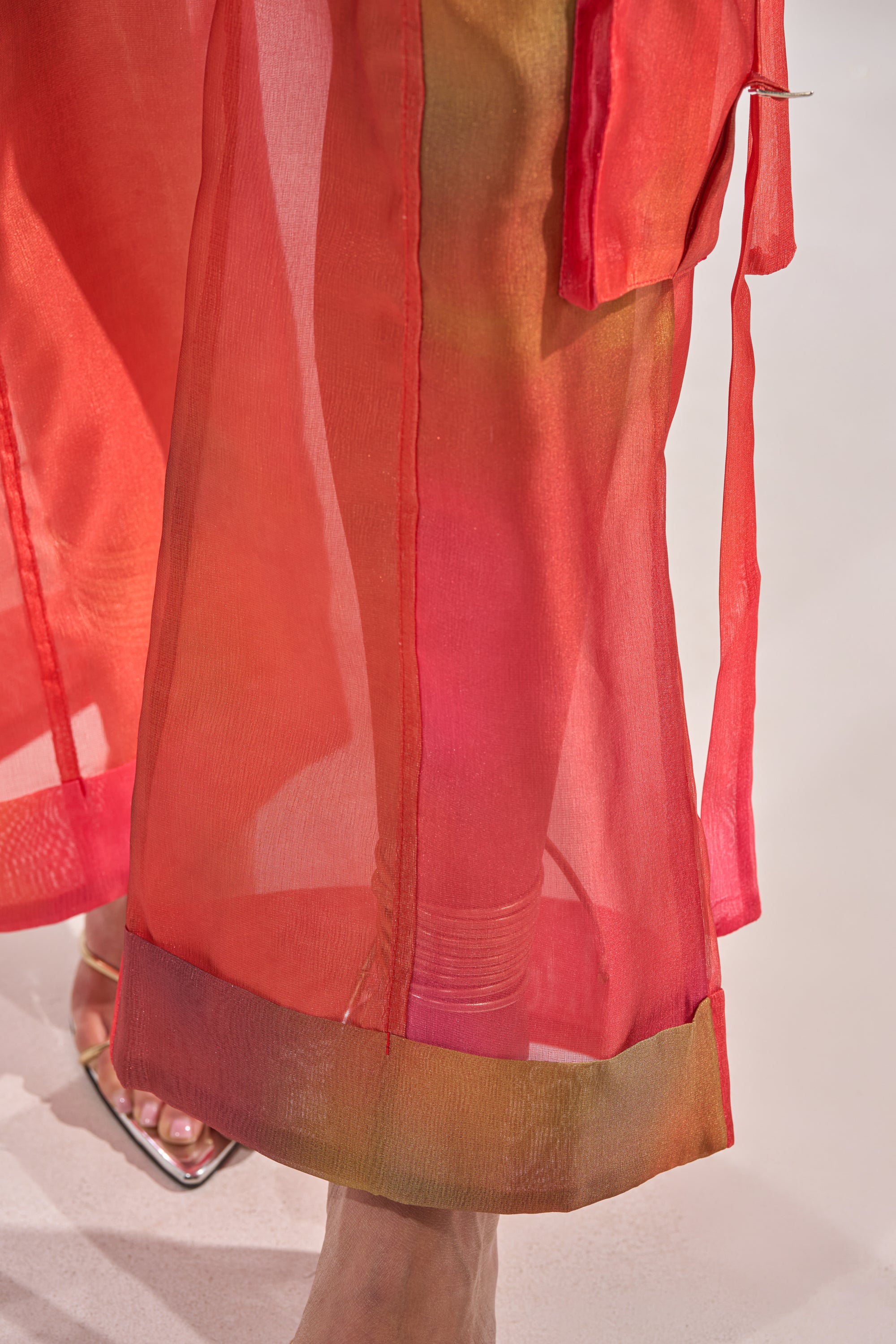A close-up of the STAINED RED LIGHTWEIGHT CARGO PANT, featuring sheer red fabric and wide-leg silhouette, with a flowy hem and layered translucent look, worn by a person showing one foot in a silver open-toe shoe. 8