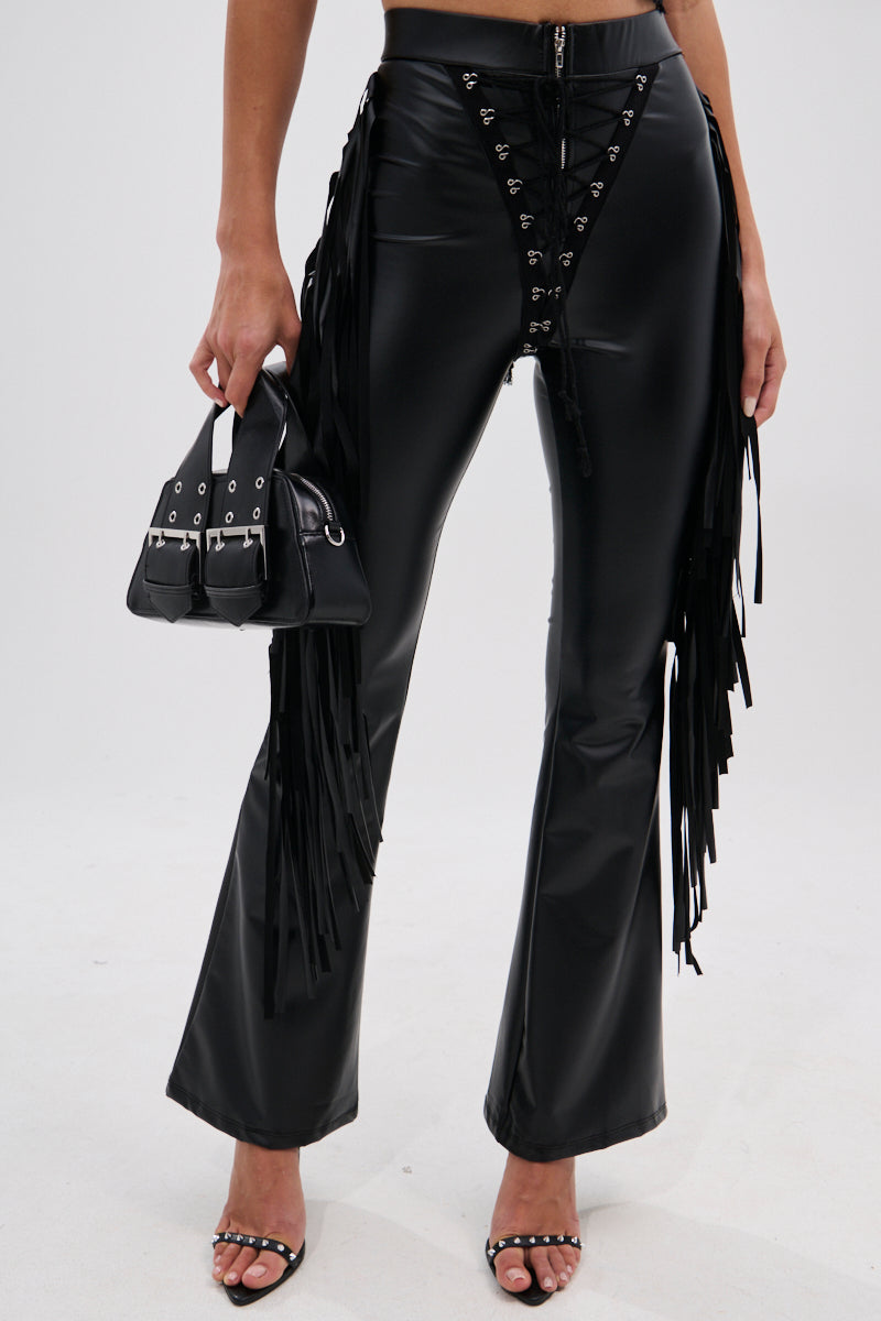 Wearing the SHOW TIME FAUX LEATHER FLARE PANT WITH LACE UP in black with fringe, paired with a small black handbag and black strappy heeled sandals for a cowgirl-chic look. The top half of the body is not shown.