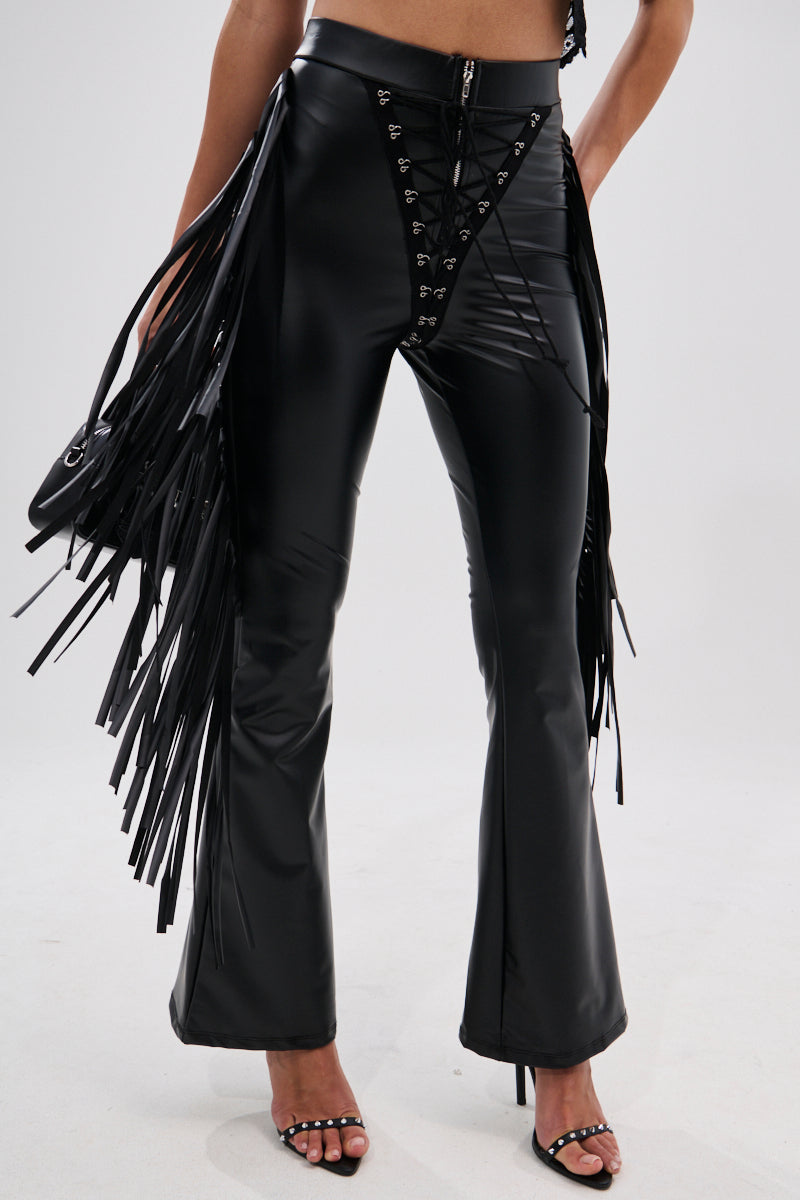 A person wears the SHOW TIME FAUX LEATHER FLARE PANT WITH LACE UP, featuring high-rise fit, lace-up front, and long black fringe on the sides with black heeled sandals, standing against a white background for a cowgirl-chic look.