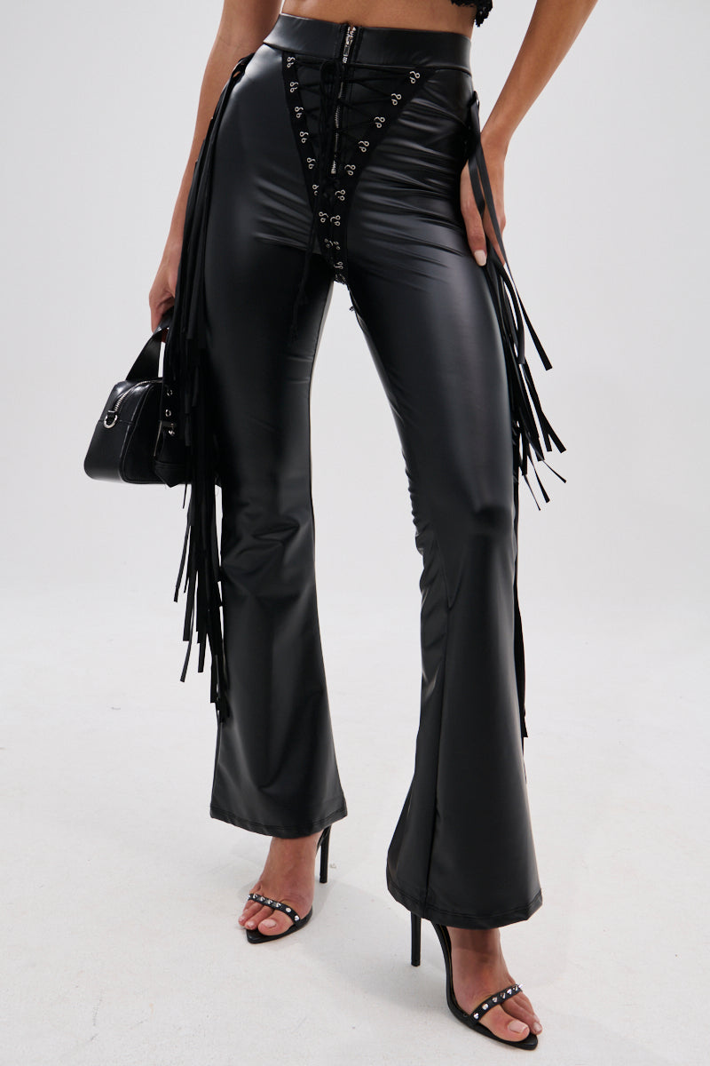 A person wears the SHOW TIME FAUX LEATHER FLARE PANT WITH LACE UP featuring high-rise fit and fringe, styled with strappy heels and a black mini handbag. The photo is cropped at the shoulders for a cowgirl-chic look.