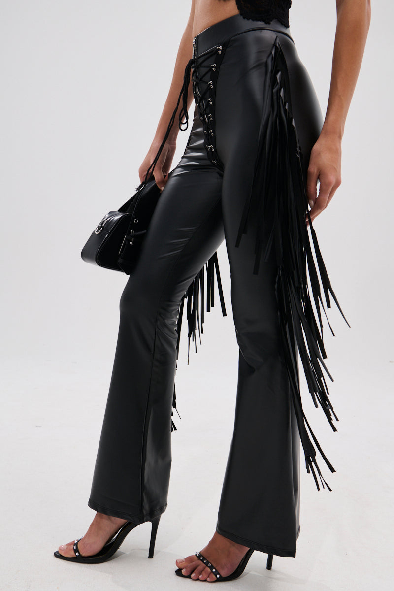 A person wears the SHOW TIME FAUX LEATHER FLARE PANT WITH LACE UP in black, featuring a high-rise fit and flare legs, styled with black high-heeled sandals and a matching handbag against a light plain background.