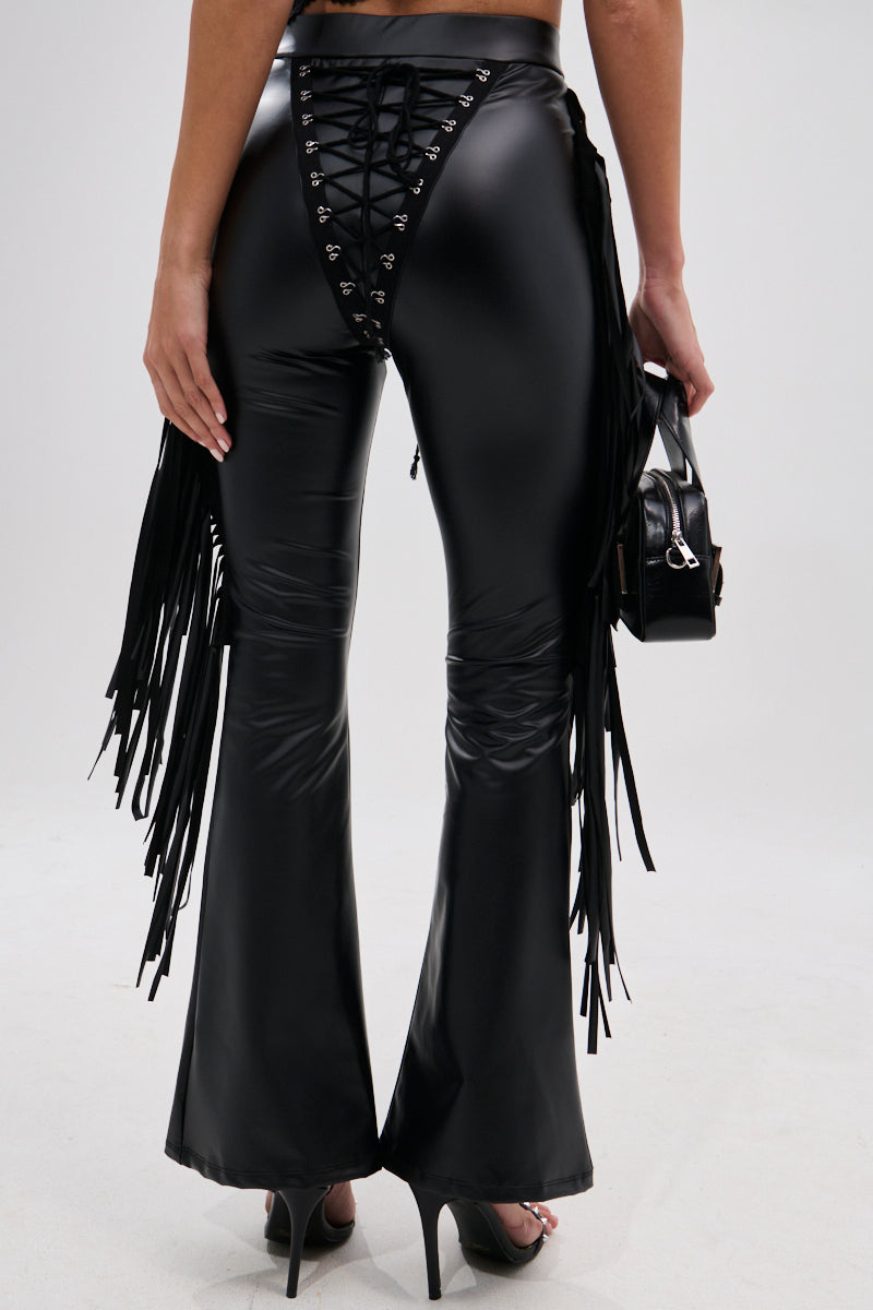 A person wears SHOW TIME FAUX LEATHER FLARE PANT WITH LACE UP—shiny black high-rise flares with side fringe—paired with a small black handbag and heels, capturing a cowgirl-chic vibe from behind against a plain background.