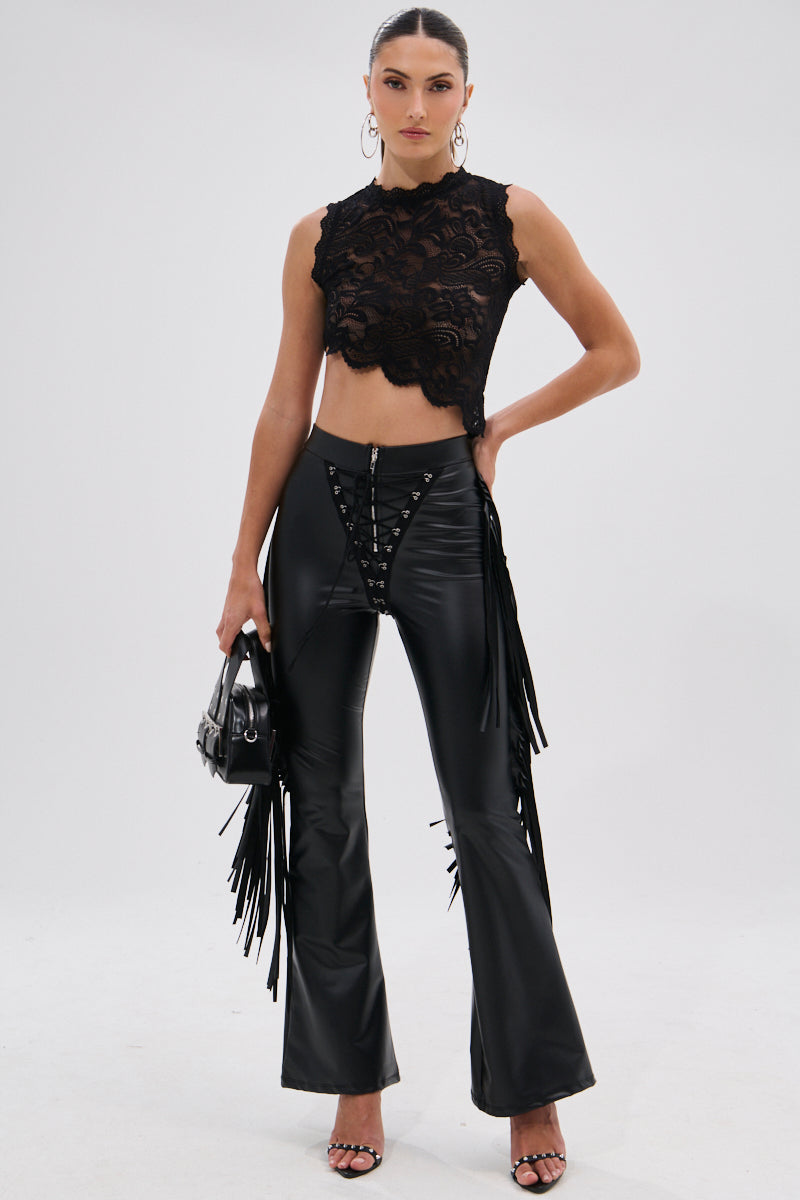 A woman wears the SHOW TIME FAUX LEATHER FLARE PANT WITH LACE UP, featuring a high-rise fit, lace-up front, and fringe sides, paired with a sleeveless black lace crop top and heels, accessorized with a small handbag for a cowgirl-chic look.