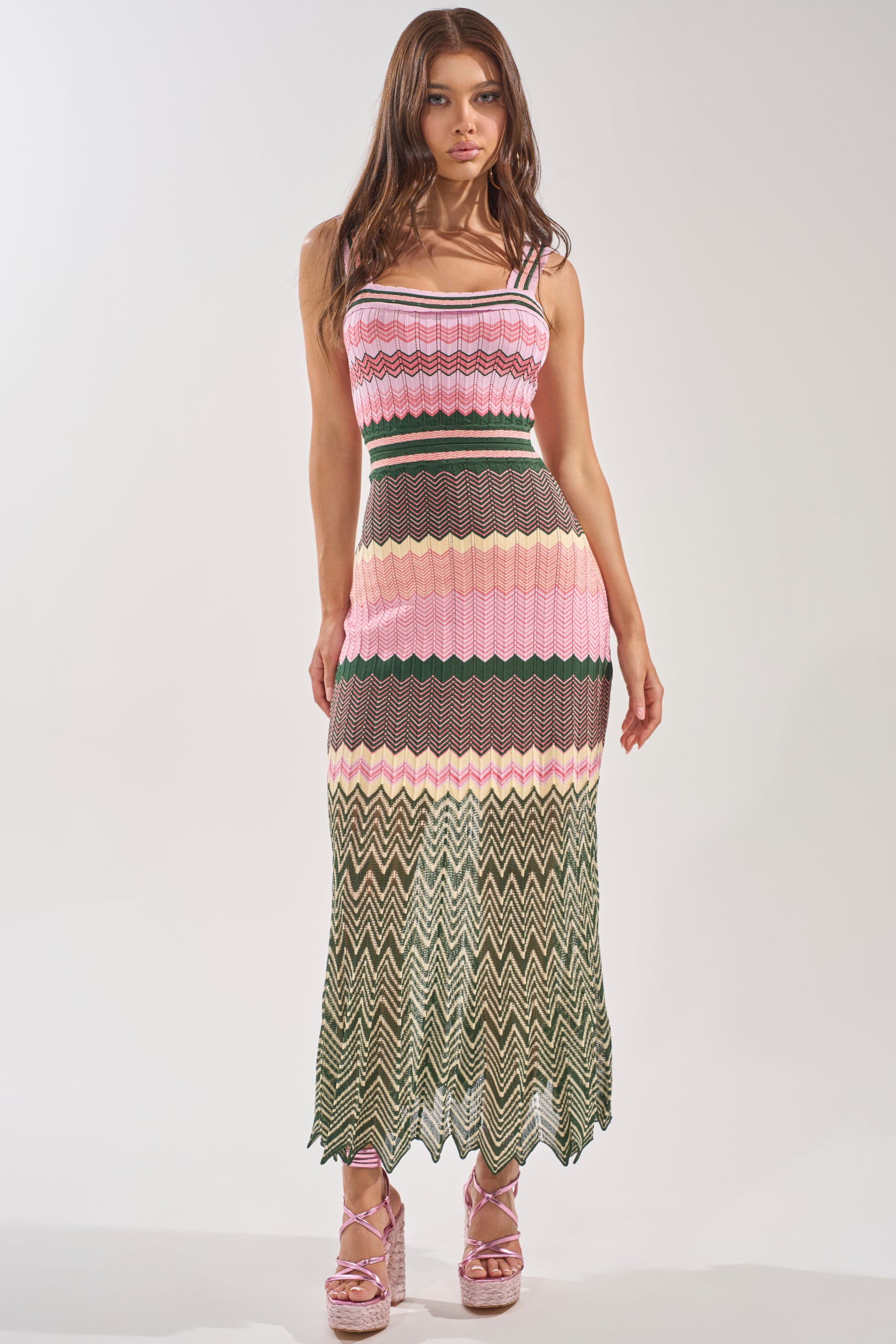 A woman models the HEAT WAVE KNIT MAXI DRESS, a sleeveless pink chevron maxi with black, yellow, and green zigzag patterns, styled with pink platform shoes against a plain light background. 0