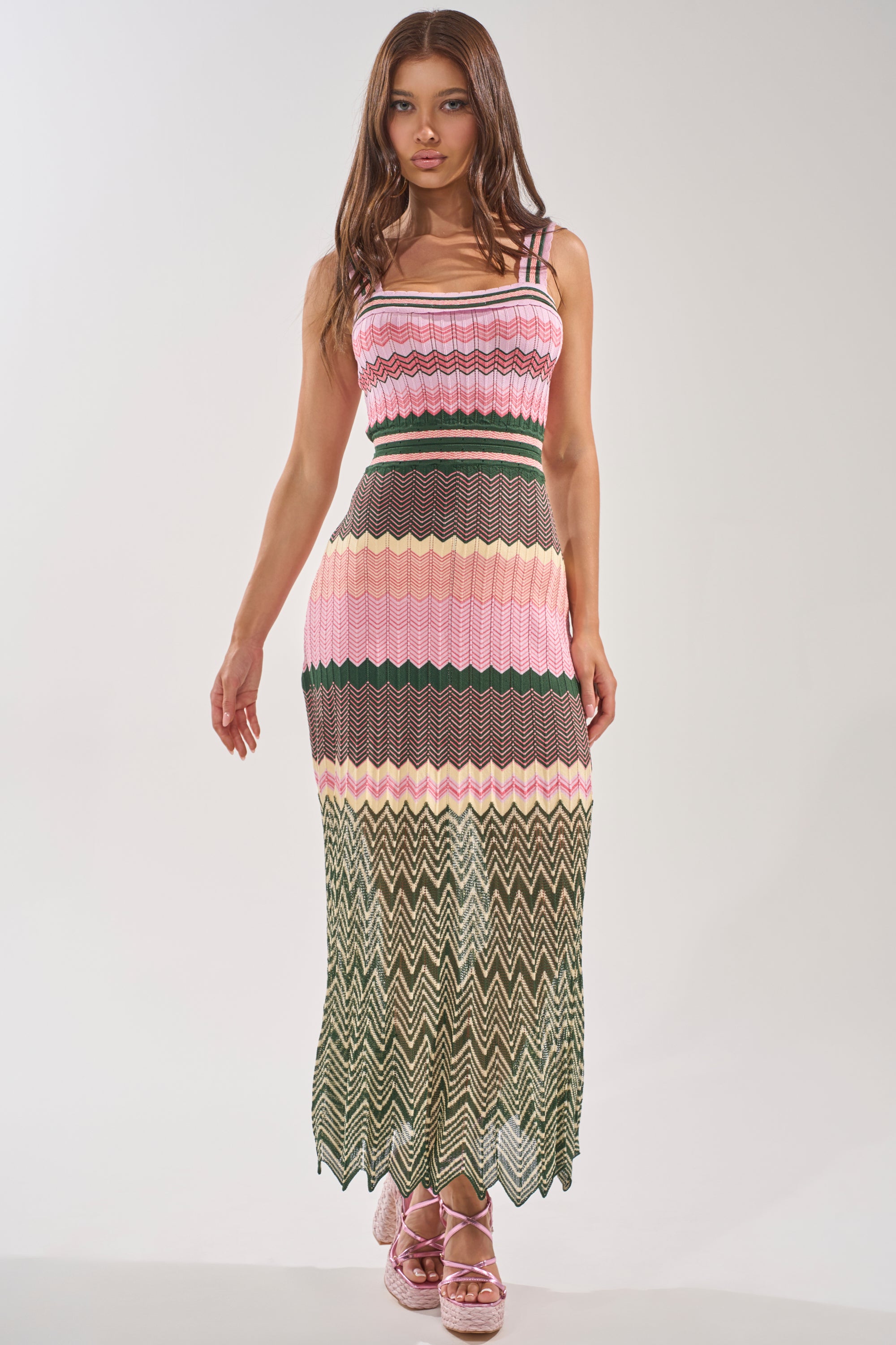 A woman models the HEAT WAVE KNIT MAXI DRESS, a color-blocked, sleeveless maxi with pink, green, and beige zigzag knit patterns. She pairs it with pink platform sandals against a plain light background—ideal for chic vacation style. 1