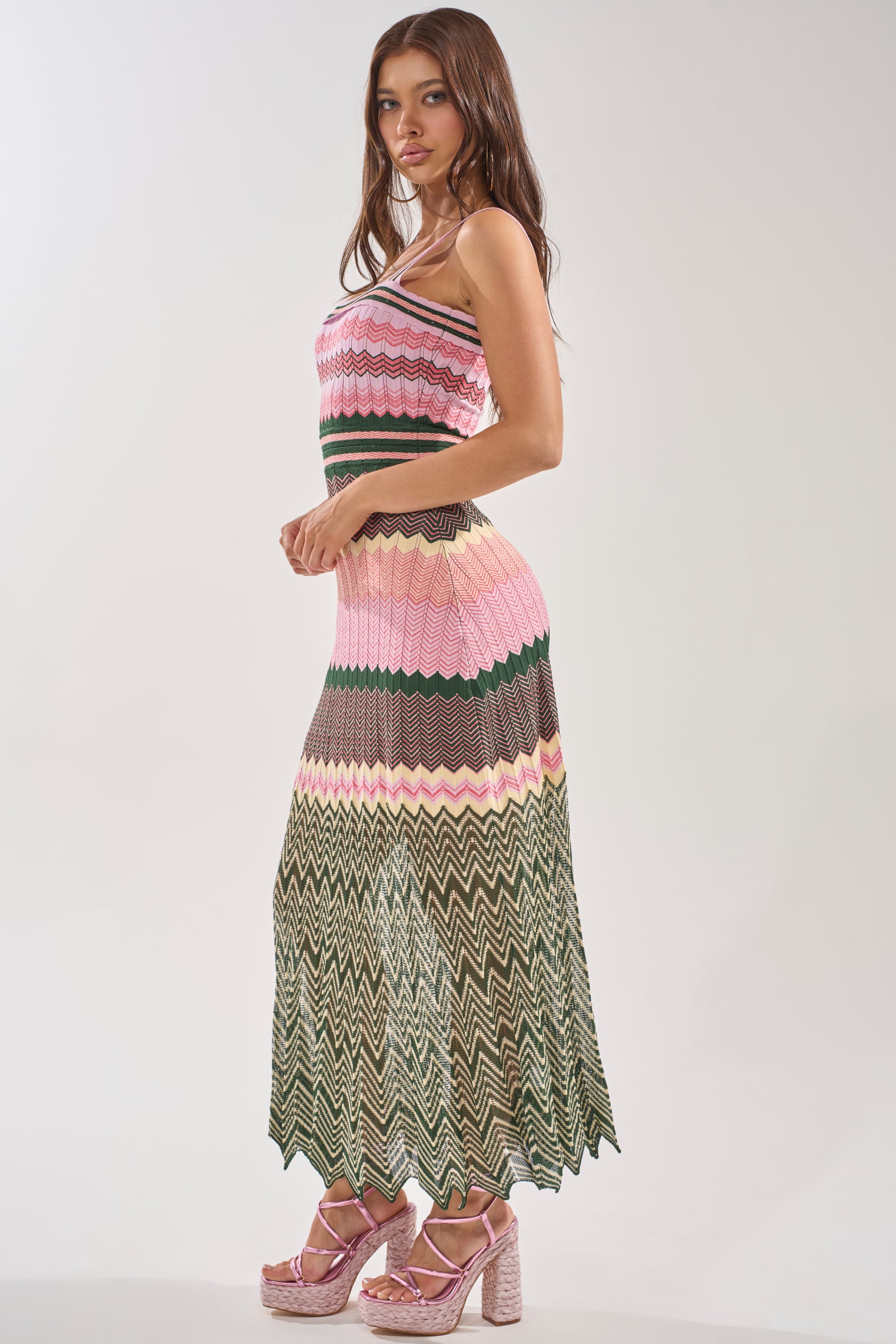 A woman stands sideways wearing the HEAT WAVE KNIT MAXI DRESS with pink, green, and beige chevron stripes, paired with pink platform heels against a plain light background. 2