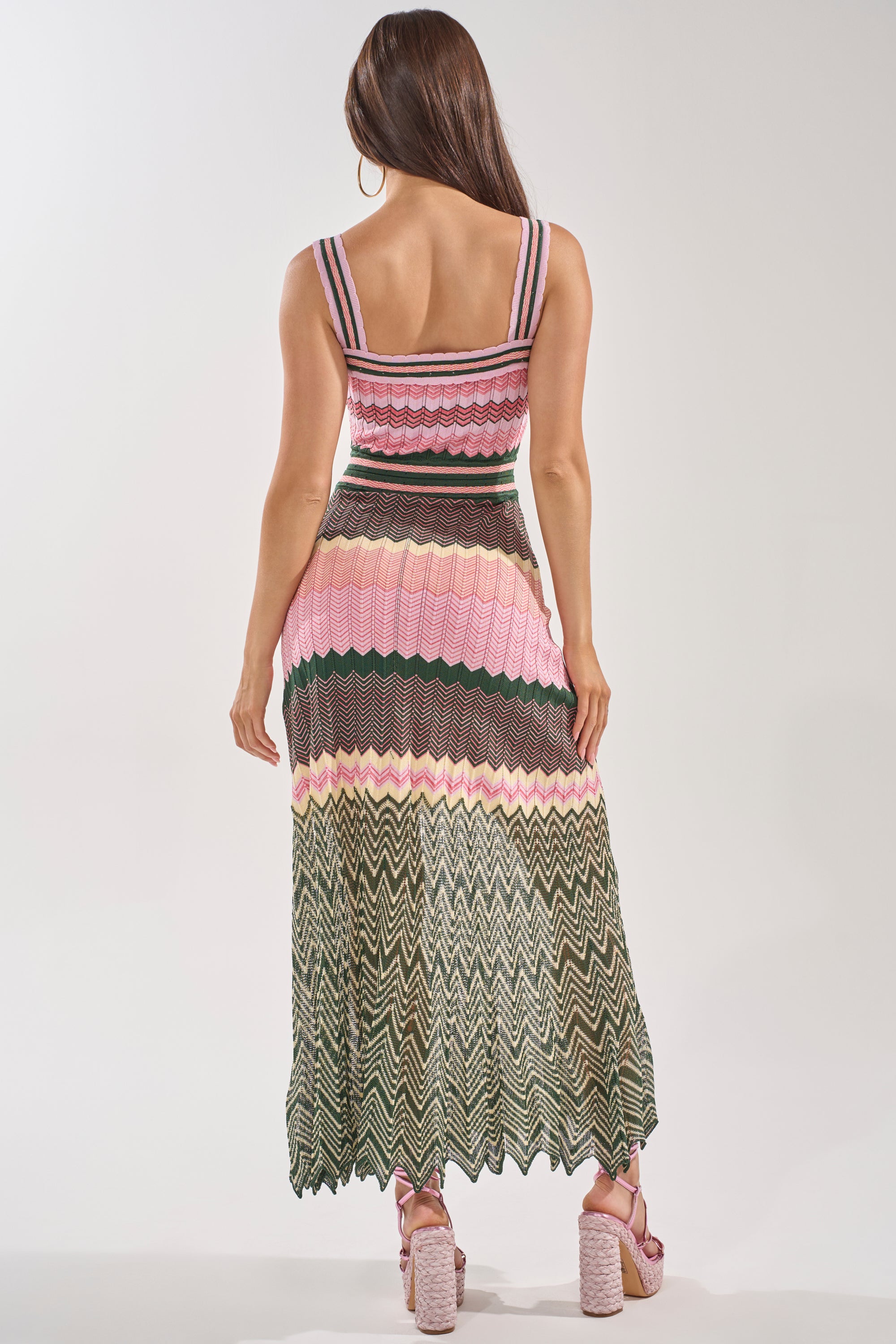 A woman with long brown hair faces away, wearing the HEAT WAVE KNIT MAXI DRESS in pink chevron with green and cream zigzags, paired with pink platform block-heel sandals—perfect for a vacation. 3