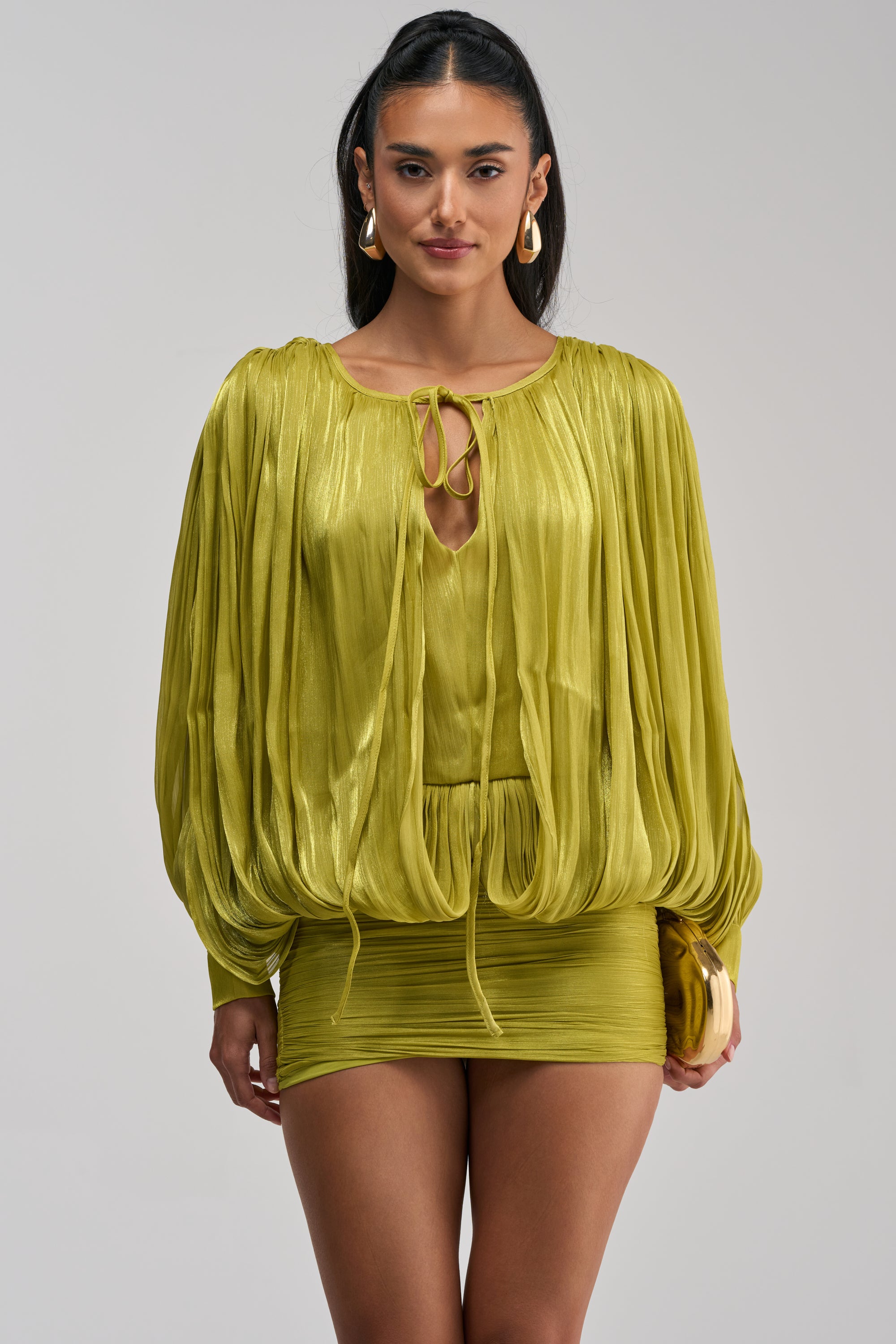 A woman models the CLASSY AND CUTE CHIFFON MINI DRESS IN GREEN—a shiny, lime green mini dress featuring long sleeves and a keyhole neckline with a ruched silhouette—standing against a plain light background.