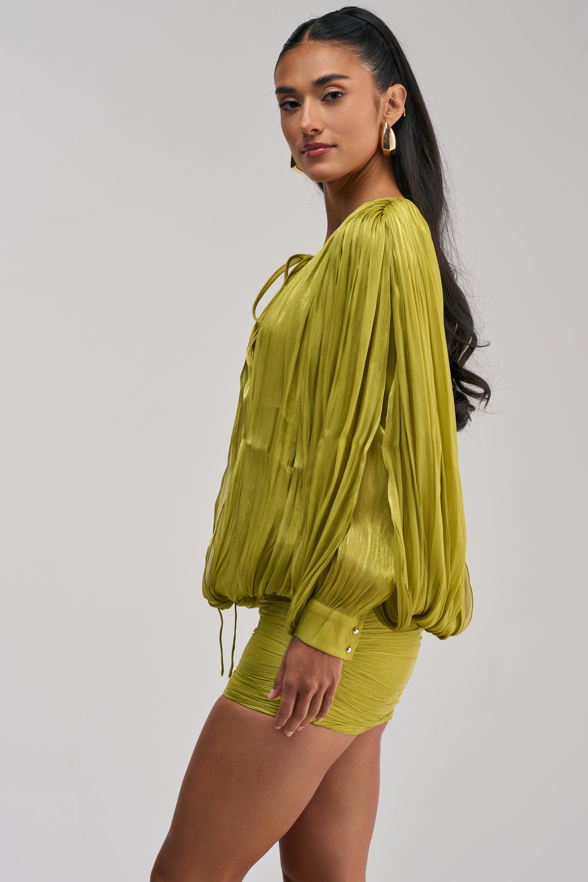 Styled with gold drop earrings, the model exudes effortless charm in the CLASSY AND CUTE CHIFFON MINI DRESS IN GREEN, featuring a draped silhouette and billowy long sleeves with a tie neckline.