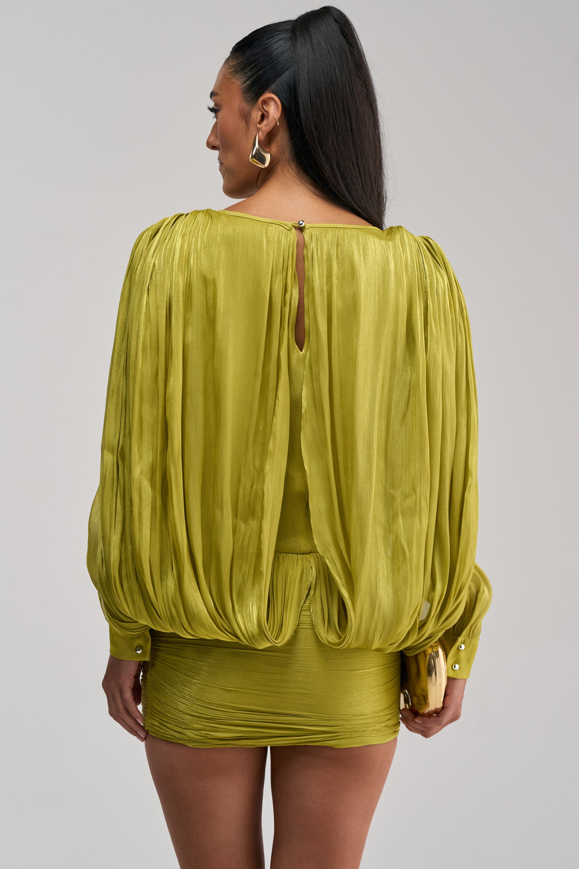 A woman with long dark hair in a high ponytail is seen from the back wearing the CLASSY AND CUTE CHIFFON MINI DRESS IN GREEN, a shiny lime green draped long sleeve mini dress. She accessorizes with a clutch and gold earrings.