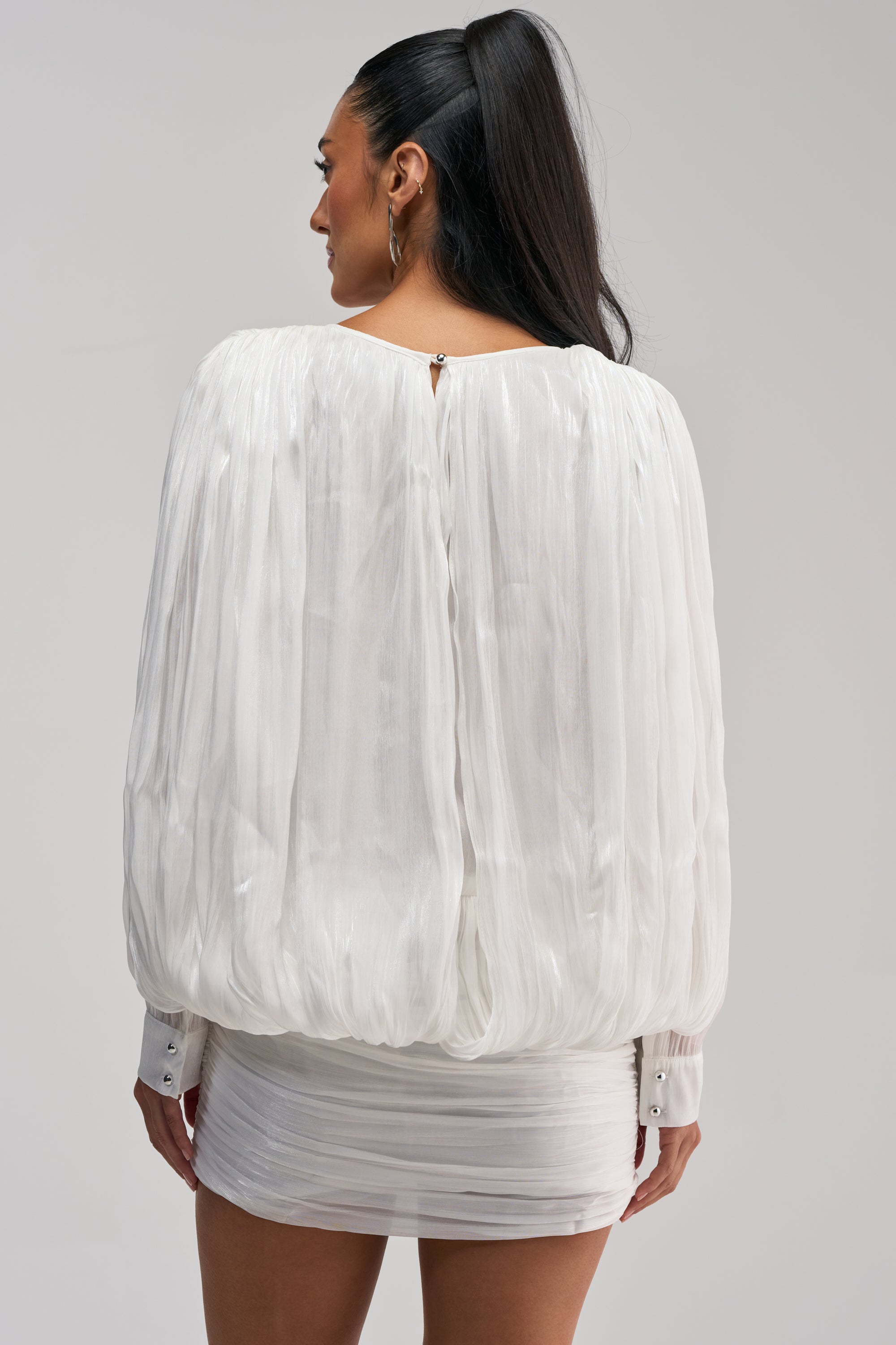 A woman with long black hair styled in a high ponytail is seen from the back against a plain background, wearing the CLASSY AND CUTE CHIFFON MINI DRESS IN WHITE—a sheer, draped long sleeve dress featuring a textured balloon silhouette.