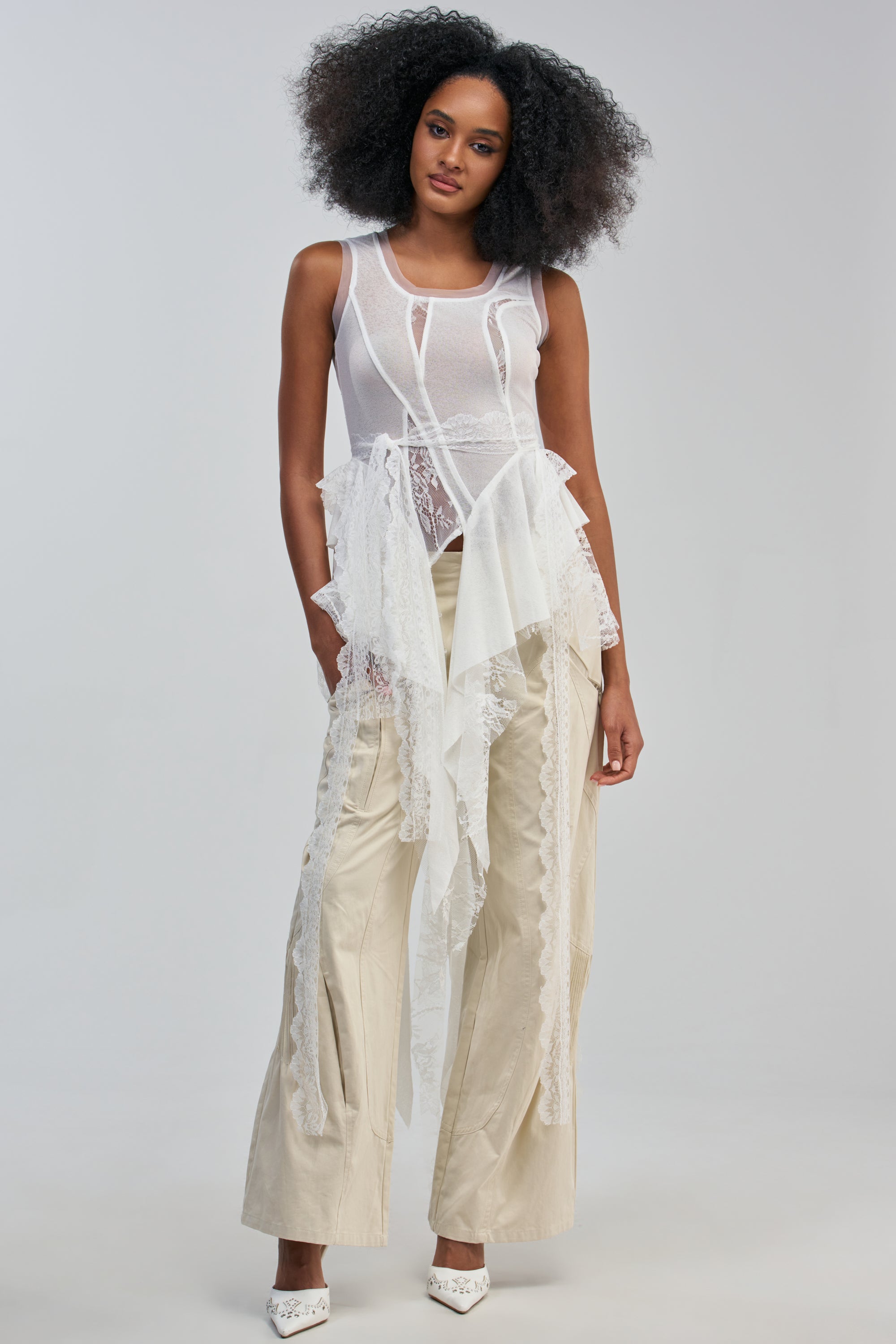 A woman with curly hair wears the ANGELIC LACE DETAIL TOP—a sheer white sleeveless top featuring lace panels and layered accents—paired with beige wide-leg pants and white heels, standing against a plain light gray background.