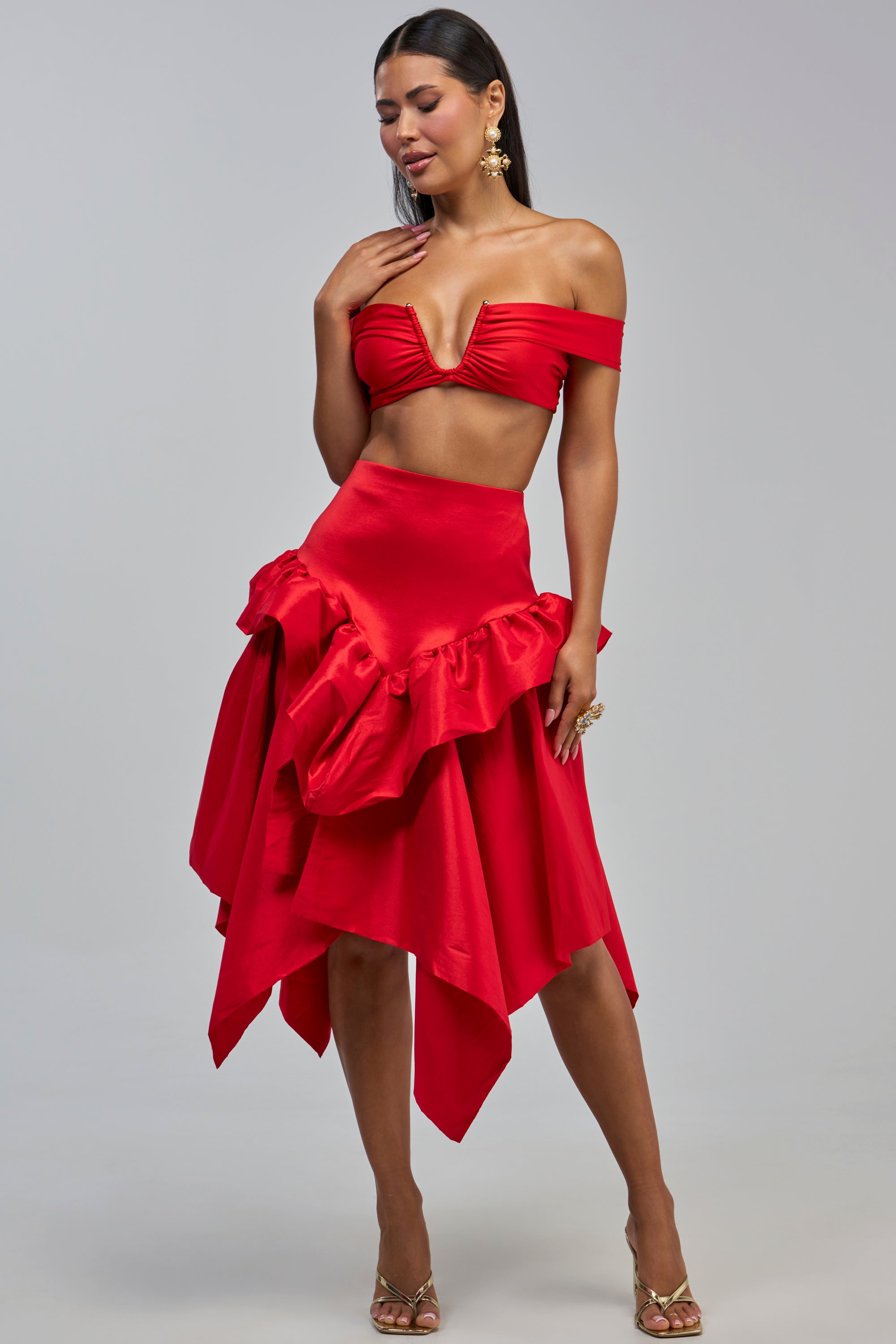 A woman with long dark hair poses against a plain gray background, wearing the striking ALL ABOUT IT MIDI SKIRT—an eye-catching red taffeta piece featuring a high waist and asymmetrical ruffles—paired with a vibrant red off-shoulder crop top, gold heels, and dangling earrings.
