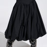 The image artistically shows the lower half of a person wearing an AKIRA Label's I FOUND YOU DROP WAIST MIDI SKIRT IN BLACK paired with pointed black boots featuring silver chain details, set against a plain background.