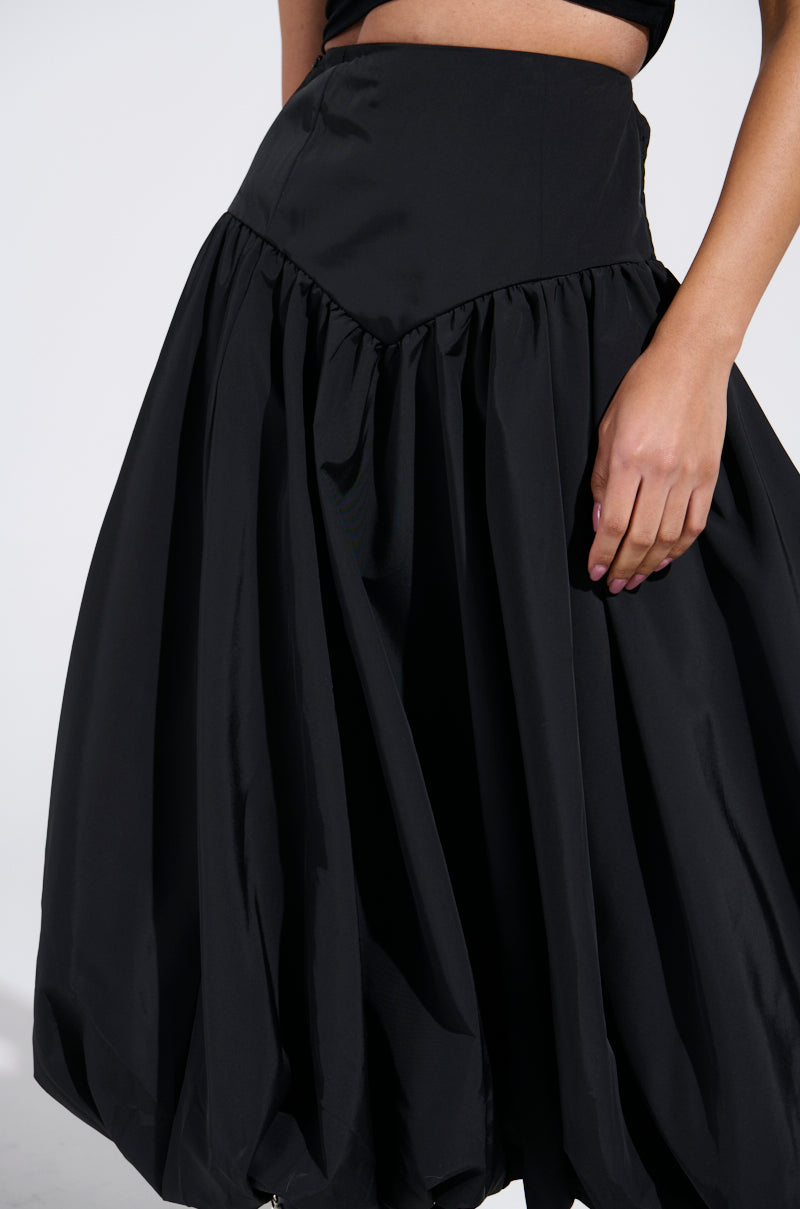 A person is wearing the AKIRA Label "I FOUND YOU DROP WAIST MIDI SKIRT IN BLACK," a voluminous skirt with a fitted waist and gathered, flowing fabric. Their hand rests on their hip against a plain, light-colored backdrop.
