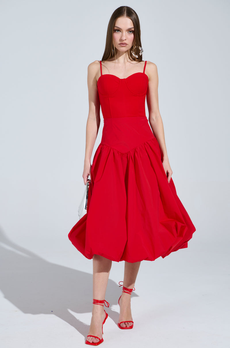 A woman with long hair wears the I FOUND YOU DROP WAIST MIDI SKIRT IN RED paired with thin straps, a full skirt, and red sandals. She holds a small white purse, standing against a plain, well-lit background that highlights the chic flow of her outfit.
