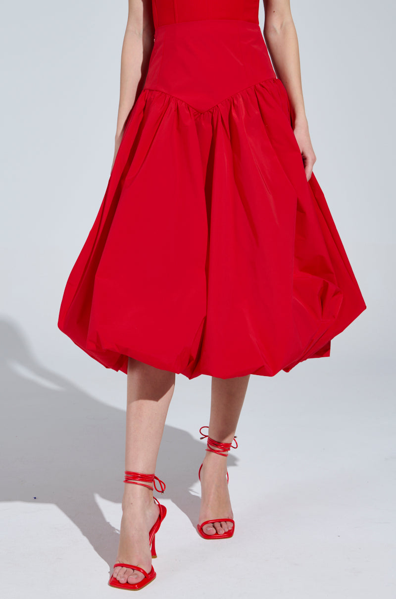 The individual dons the "I Found You Drop Waist Midi Skirt in Red," characterized by its voluminous flair, paired with red strappy high heels. The photo showcases the sleek outfit against a plain background, highlighting the skirt's and shoes' elegance.