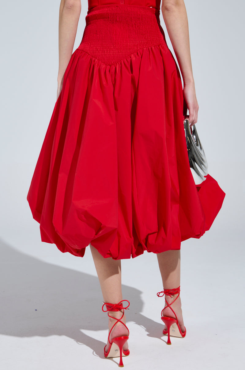 A person wearing the I FOUND YOU DROP WAIST MIDI SKIRT IN RED with matching high-heeled lace-up shoes is holding a silver handbag. The skirt features a gathered waist and balloon shape against a light gray background.