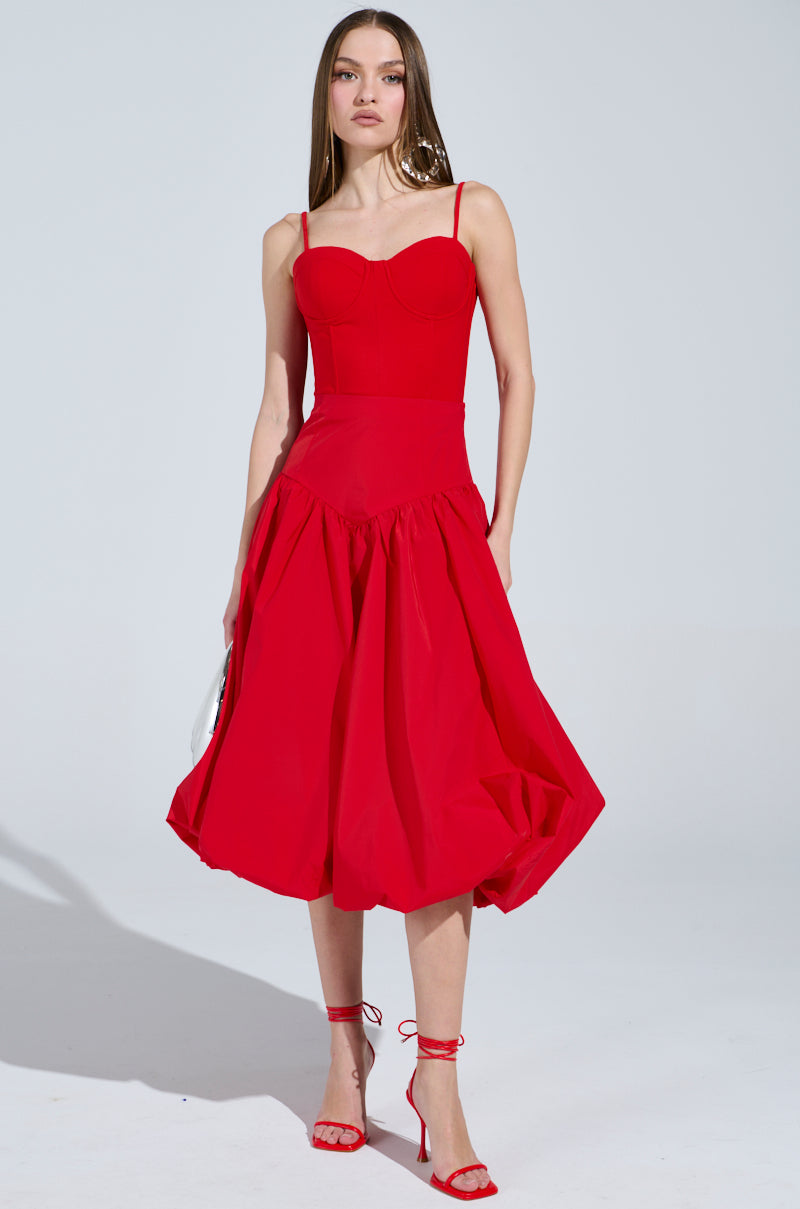 A woman with long brown hair is wearing a sleeveless red dress featuring I Found You Drop Waist Midi Skirt. Her look is completed with red high-heeled sandals against a plain, light-colored background.