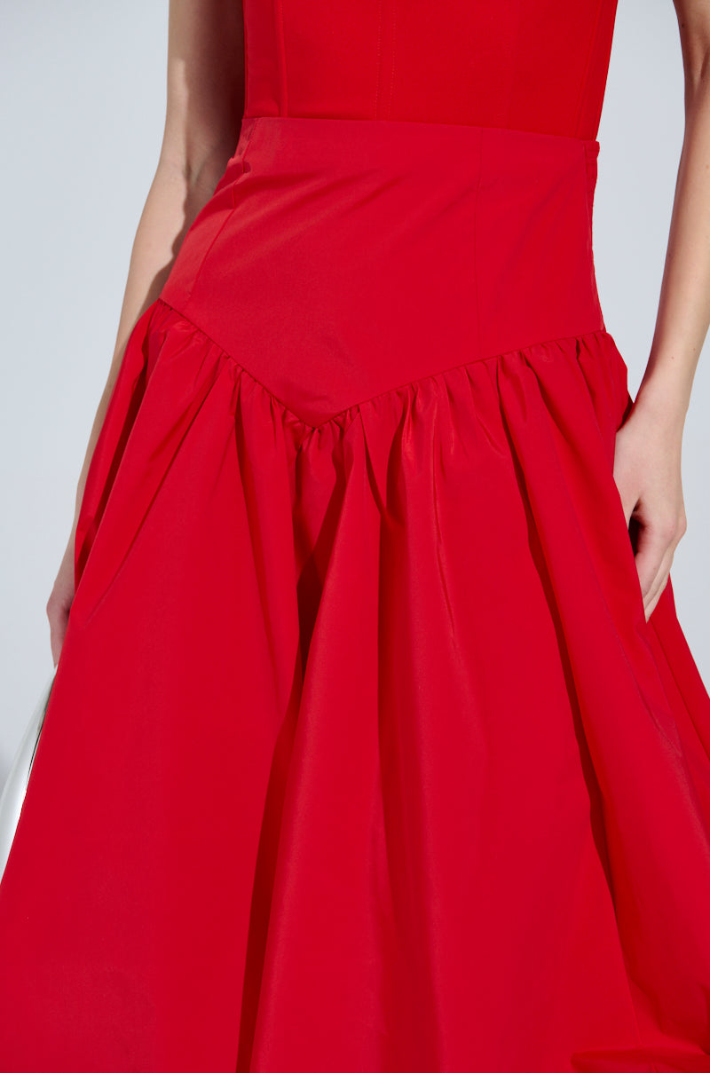 A person wears the "I Found You Drop Waist Midi Skirt in Red," featuring a gathered waist that flows into a voluminous skirt. The bright red fabric stands out against a neutral gray background, with only part of the arm visible.