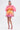 A woman stands wearing a vibrant outfit featuring the Shelby Ombre Mini Skirt and matching blouse, paired with pink heels. The pleated, fan-shaped pink and orange top pops against the plain white background.