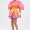 A woman stands wearing a vibrant outfit featuring the Shelby Ombre Mini Skirt and matching blouse, paired with pink heels. The pleated, fan-shaped pink and orange top pops against the plain white background.