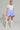 A person stands confidently wearing a white short-sleeved blouse and the DREAMERS BUBBLE SKIRT in blue stripes with ruffles. They hold a white purse and wear tall white lace-up boots, accessorized with hoop earrings against a plain light-colored background.
