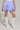 A person dressed in the DREAMERS BUBBLE SKIRT, featuring a striped blue design with ruched details and a high-rise fit, holds a white purse and wears white knee-high lace-up boots against a plain background that highlights the chic ensemble.