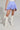 A person wearing the DREAMERS BUBBLE SKIRT, featuring blue and white stripes with gathered and ruched details, paired with a high-rise fit. They sport white lace-up knee-high boots and hold a white bag against a plain, light gray background.