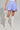 A person wearing the DREAMERS BUBBLE SKIRT, featuring blue and white stripes with drawstring details and a tulle inner-lining, is styled with knee-high white lace-up boots and holds a small white purse. The background is plain and neutral.