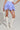A person stands wearing a white corset top paired with the DREAMERS BUBBLE SKIRT, which features a blue and white pattern and high-rise ruching. They complement their outfit with white knee-high lace-up boots and carry a small white handbag. The background is plain gray.