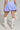 A person stands wearing the DREAMERS BUBBLE SKIRT, a blue and white skirt with a tulle inner-lining, paired with a white top and tall white boots. They hold a white handbag in their left hand against a plain background.