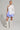 A person poses confidently in a fashion-forward outfit, featuring a white one-sleeved top, the DREAMERS BUBBLE SKIRT in blue and white stripes with gathered details, and knee-high white lace-up boots. They complement their look with hoop earrings and have their hair styled back.