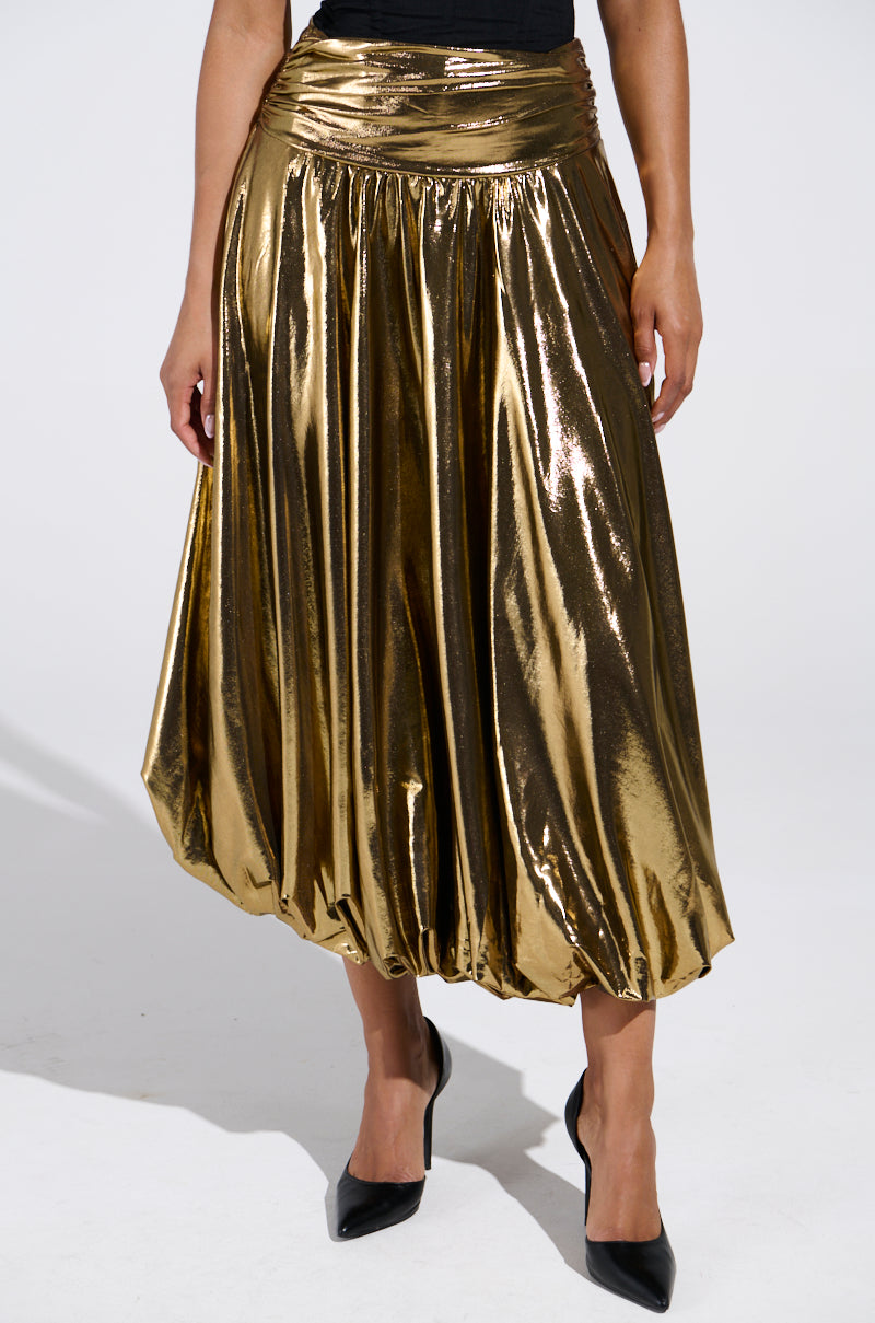 Wearing the MONEY MAKER METALLIC MIDI SKIRT, gathered at the waist and flowing loose to the calves, with black pointed high heels, a person stands against a plain background.