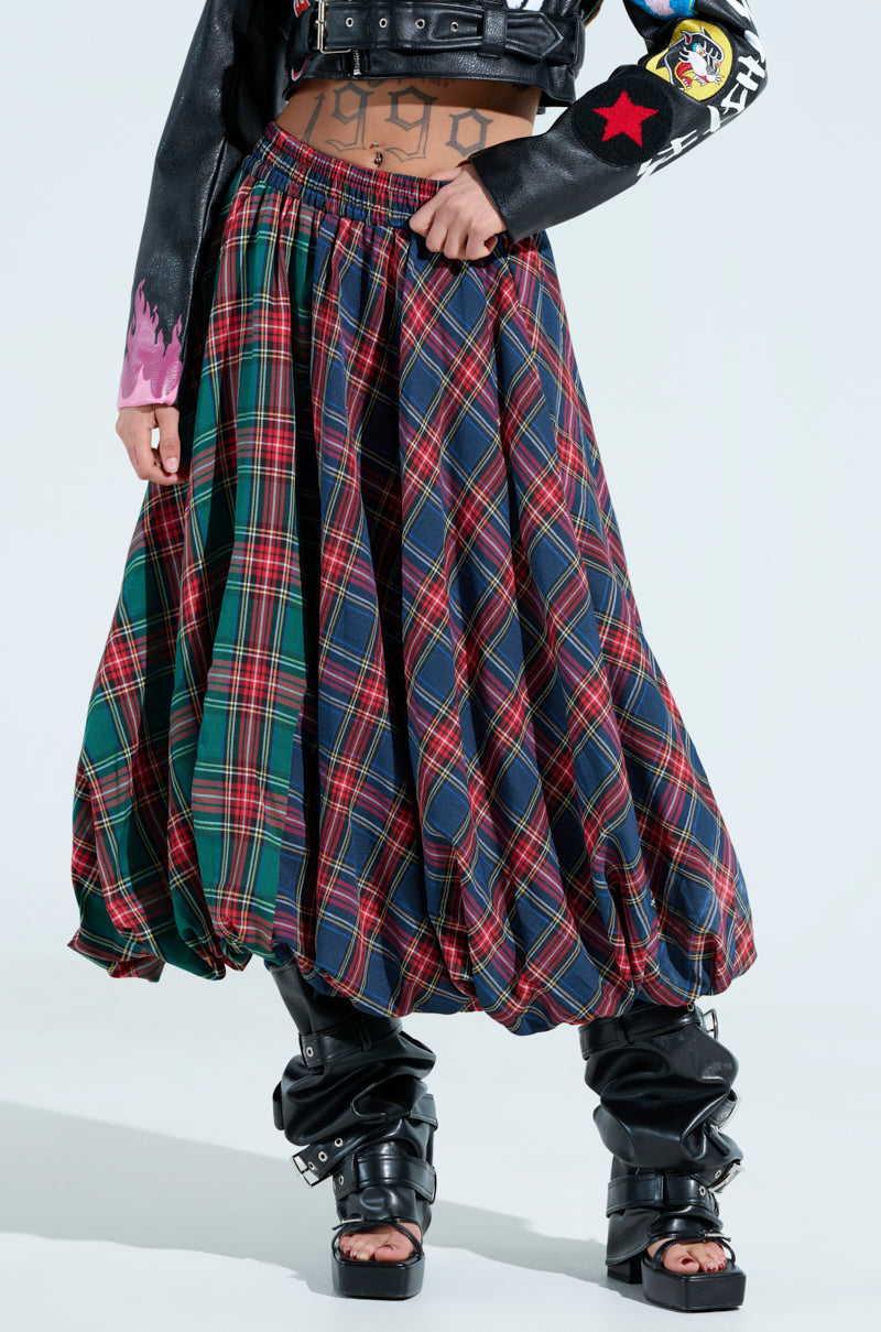 Wearing the vibrant MOVEMENTS PLAID MIDI SKIRT with a black leather jacket featuring patches, paired with edgy black platform boots against a light, neutral backdrop.