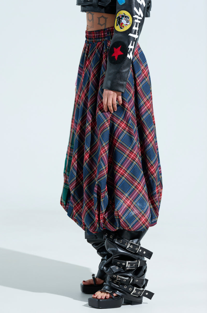 A person is wearing the MOVEMENTS PLAID MIDI SKIRT with black buckle boots. Their visible right arm sports a black sleeve decorated with various patches, all set against a plain white background that emphasizes the tartan plaid design of the skirt.