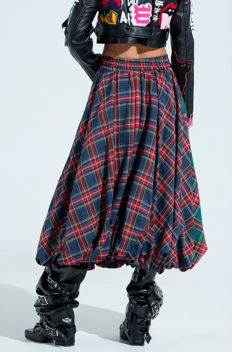 The image showcases the lower half of a person facing away, dressed in a punk-inspired style wearing the MOVEMENTS PLAID MIDI SKIRT paired with a black leather jacket adorned with colorful designs and black boots. Bold patterns and textures define this edgy look.