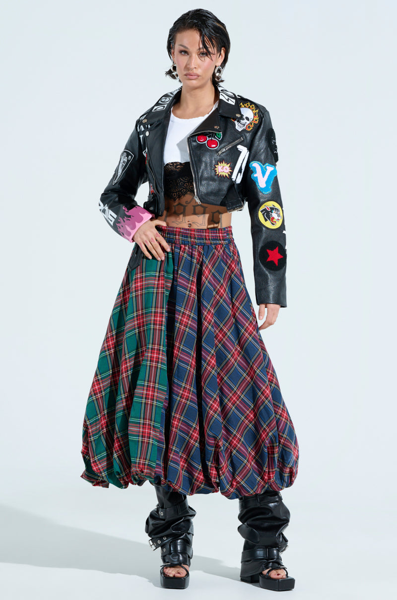 Against a plain background, a person stands confidently, dressed in a black leather jacket with colorful patches over a white shirt and black lace top. Their ensemble features the MOVEMENTS PLAID MIDI SKIRT, showcasing vibrant tartan patterns, and sleek black boots complementing their short hair.