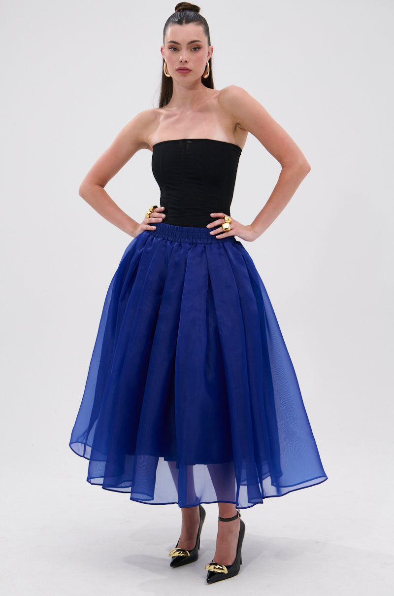 A woman stands with hands on hips in a black strapless top, bold gold jewelry, black pointed heels, and the STAY CLOSE TO ME MIDI SKIRT IN BLUE, featuring a voluminous A-line silhouette, against a plain white background. 0