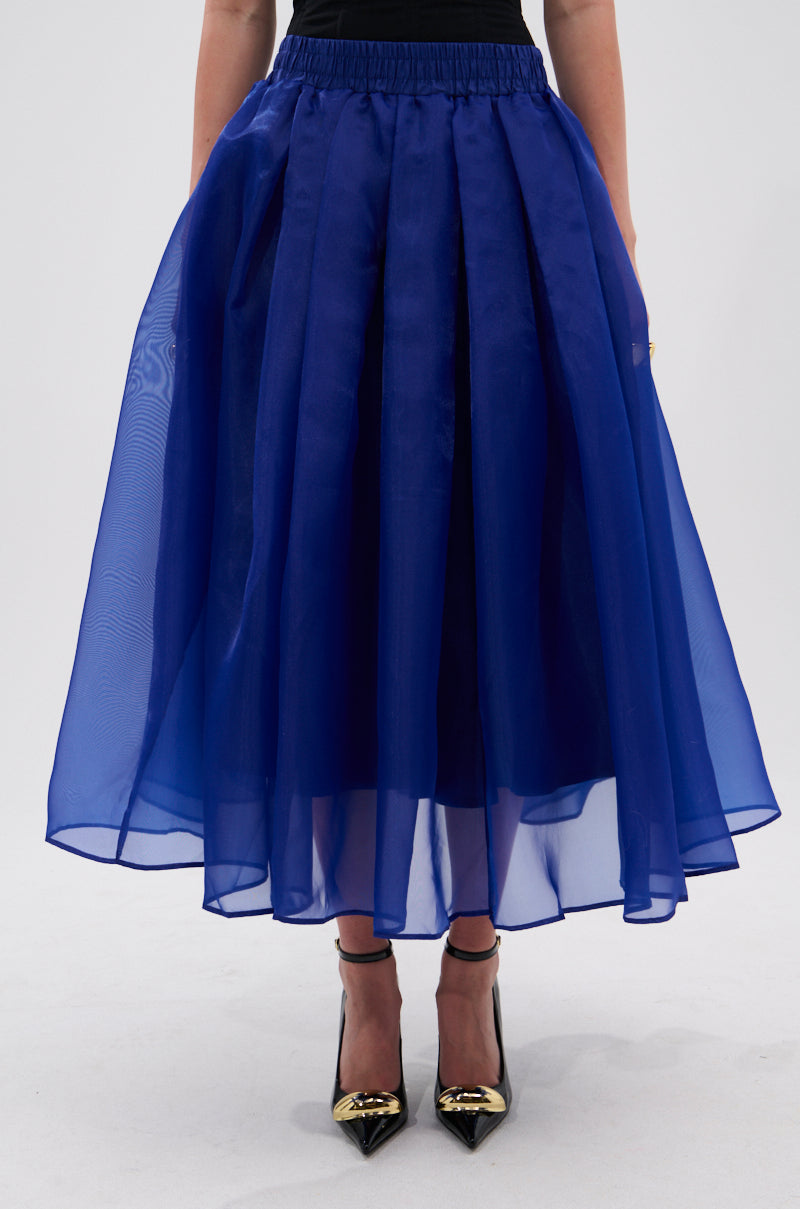 A person wears the STAY CLOSE TO ME MIDI SKIRT IN BLUE, featuring a sheer chiffon A-line silhouette, paired with a black top and black pointed-toe heels with gold accents, standing against a plain light backdrop. 1
